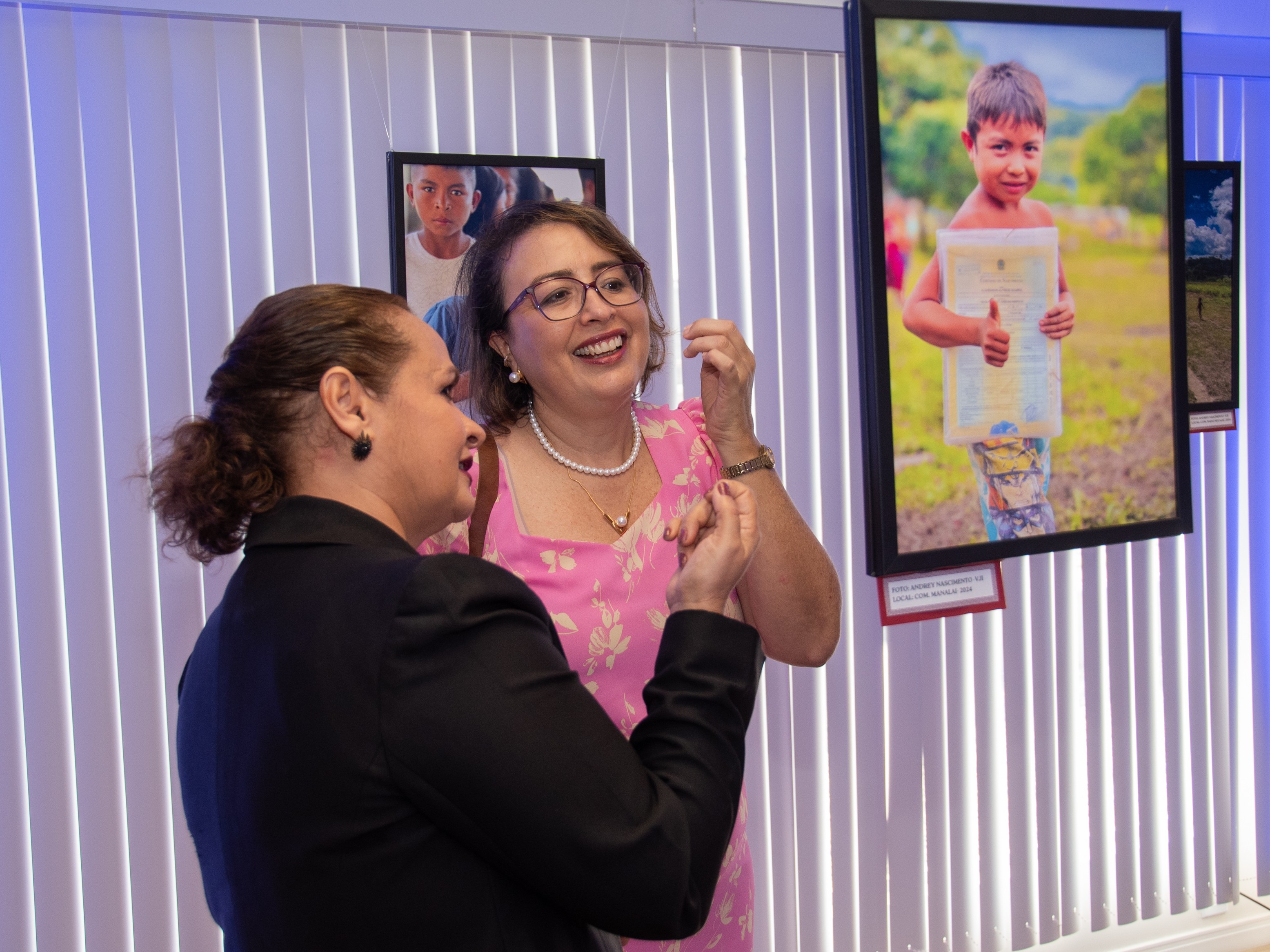 Imagem mostra duas mulheres conversando sorridentes em frente a fotografias expostas em uma galeria. Uma delas veste blazer preto e tem o cabelo preso; a outra usa vestido rosa estampado, óculos e colar de pérolas. Ao fundo, uma grande foto mostra um menino sorridente segurando um documento e fazendo sinal de positivo com o polegar. As imagens estão fixadas em molduras pretas suspensas, diante de persianas brancas iluminadas com luz azul.