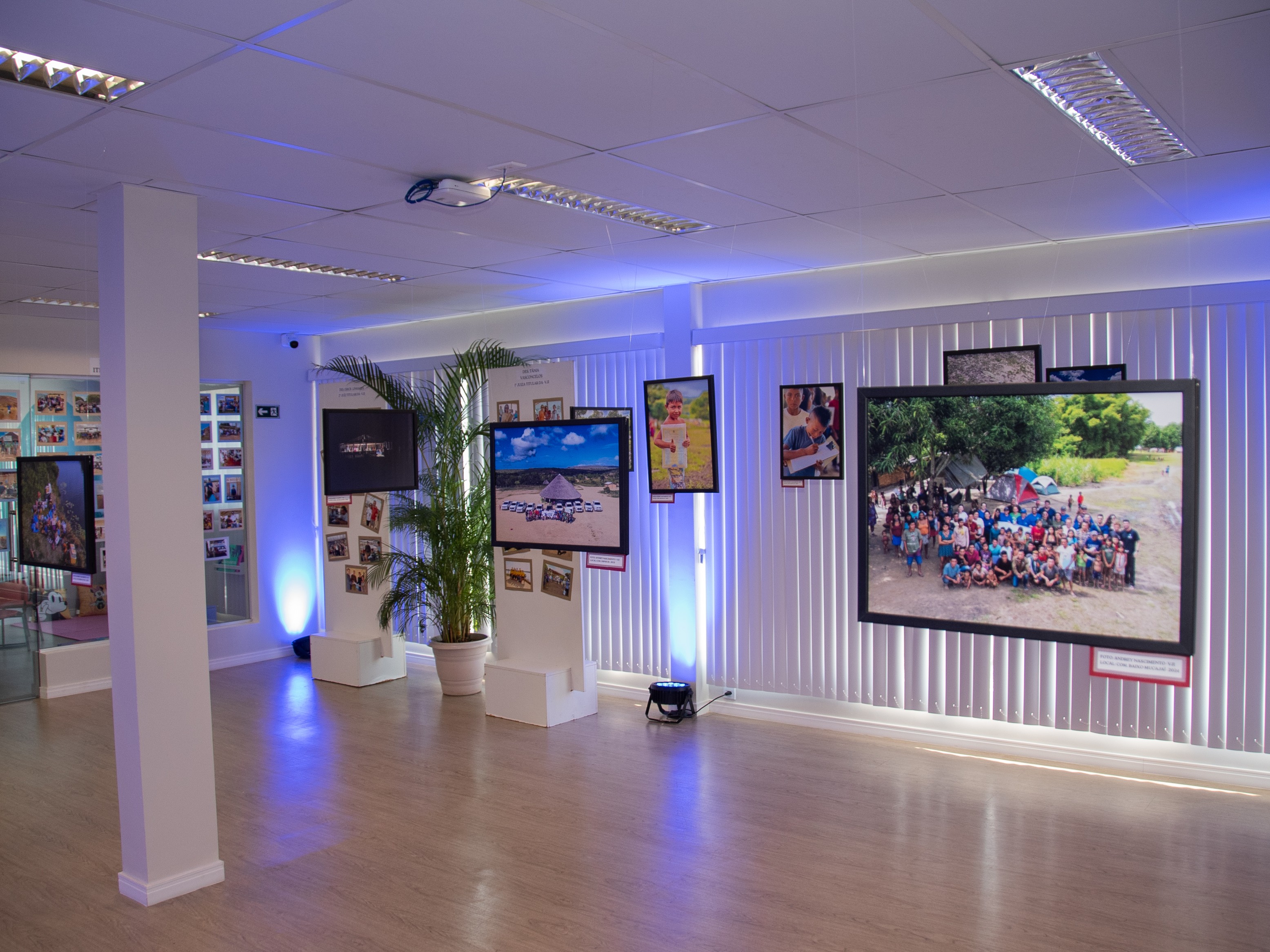  Imagem mostra um ambiente iluminado em tons de azul e branco que exibe uma mostra fotográfica com imagens de comunidades, crianças e paisagens. As fotografias, dispostas em grandes molduras suspensas e painéis ao redor da sala, destacam momentos de interação social, cenas do cotidiano e registros coletivos.