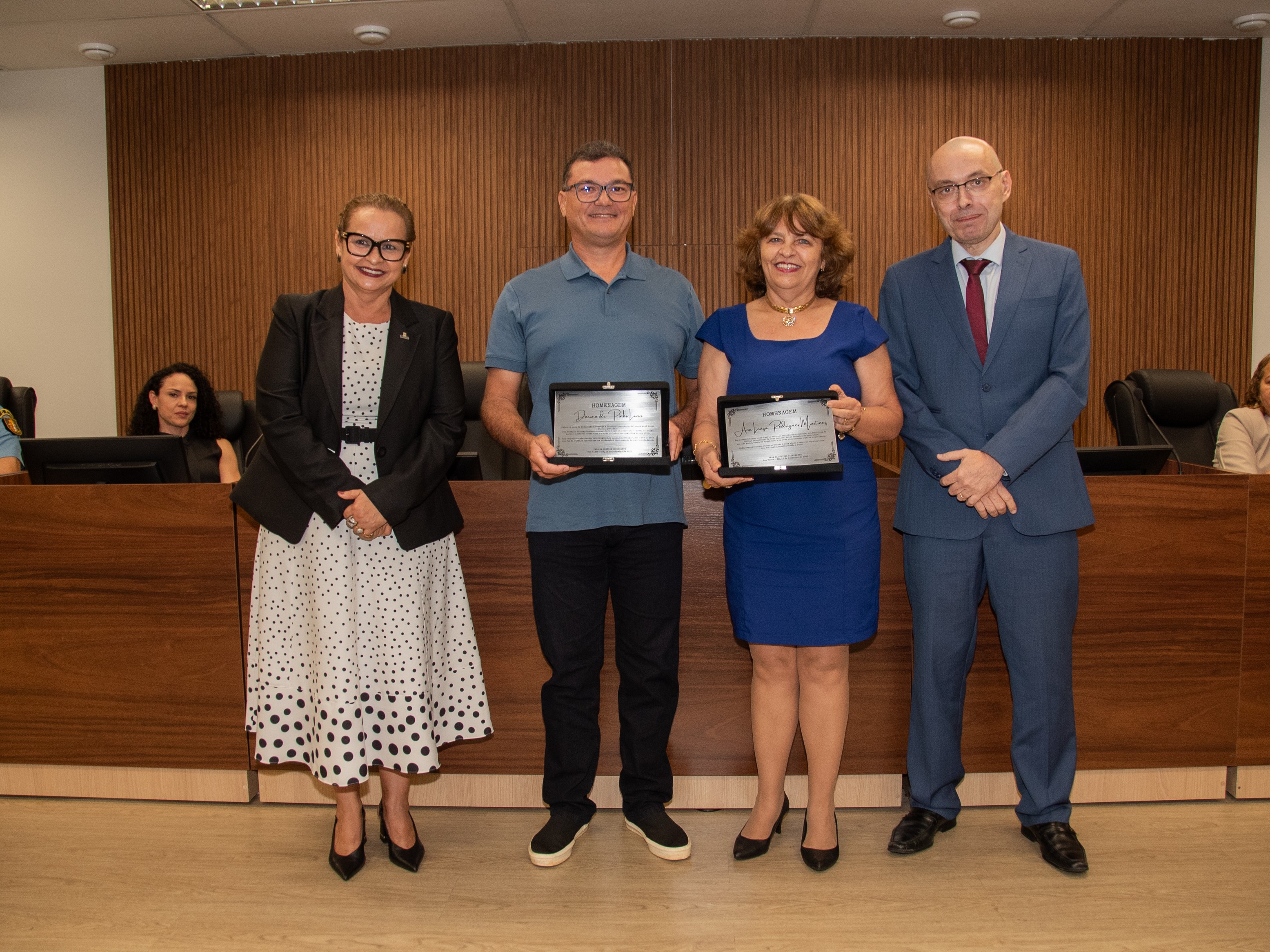 Imagem mostra quatro pessoas posando para foto em frente a uma mesa de autoridades em ambiente formal. Duas delas, ao centro, seguram placas de homenagem: um homem de óculos, camisa polo azul e calça escura, e uma mulher com vestido azul. Ao lado esquerdo, uma mulher de vestido branco com bolinhas pretas e blazer preto sorri para a câmera; à direita, um homem de terno azul e gravata vermelha olha para frente. Ao fundo, outras pessoas acompanham sentadas.