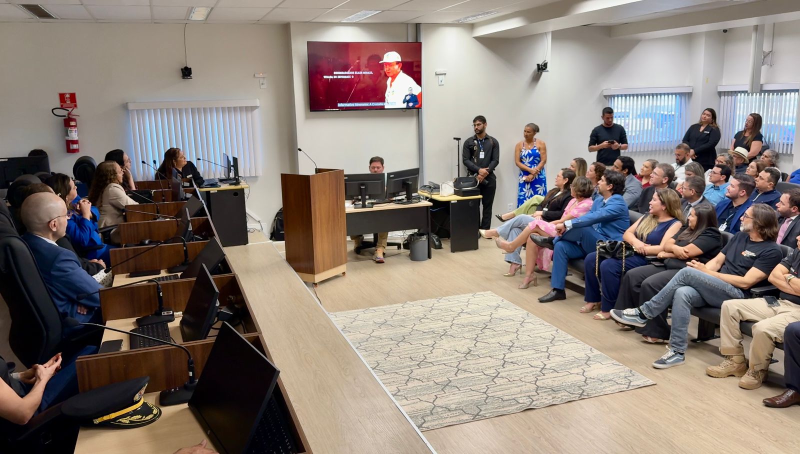 Imagem colorida mostra um auditório lotado durante uma apresentação institucional. À esquerda, pessoas estão sentadas nas bancadas do plenário, com notebooks à frente, formando uma fileira voltada para o púlpito. No centro, um homem opera dois computadores sobre uma mesa. No lado direito da imagem, o público assiste atentamente a um vídeo exibido em uma tela grande na parede, que mostra um homem