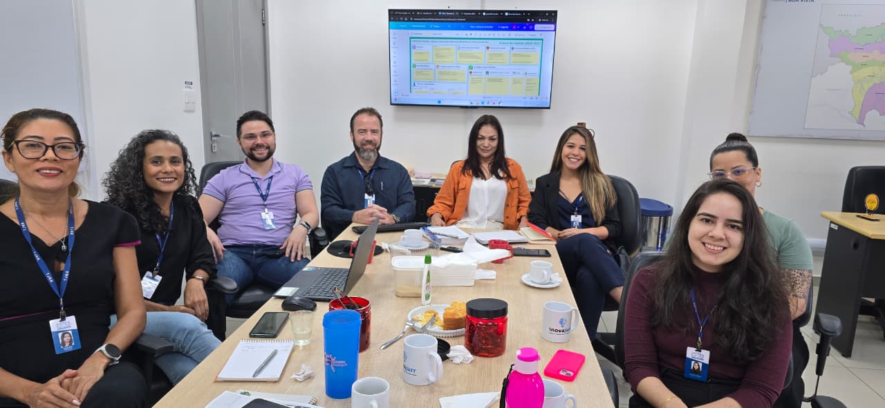 Imagem de um grupo de nove pessoas sentadas em torno de uma mesa de reunião. Sobre a mesa, há notebooks, canecas, copos, garrafas de água e alguns potes com lanches. Todos olham para a câmera e sorriem. Ao fundo, uma televisão presa na parede exibe uma tela com tabelas e gráficos. O ambiente é uma sala clara, com paredes brancas.