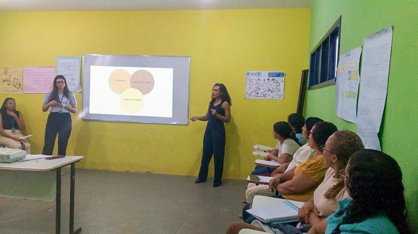  Imagem de uma sala de aula. Duas mulheres estão em pé em frente a um quadro branco, fazendo uma apresentação. No quadro, é projetada uma imagem com três círculos coloridos que se sobrepõem. À direita, várias pessoas estão sentadas em carteiras, observando e anotando. Há cartazes colados nas paredes.