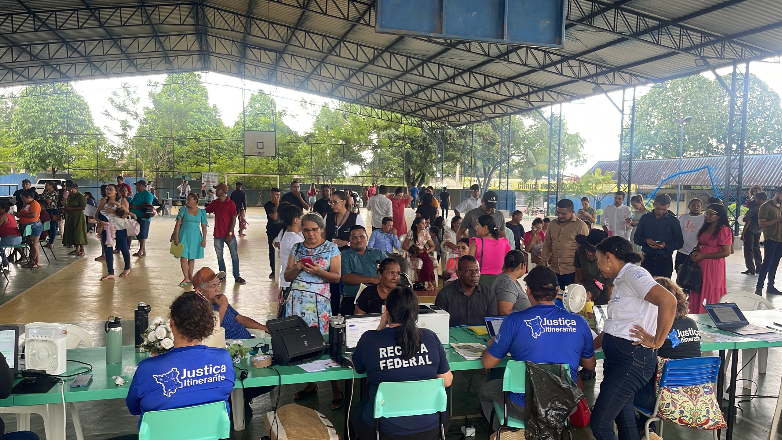  Imagem colorida mostra um ginásio coberto e bem iluminado onde ocorre um evento de atendimento ao público. Várias pessoas, algumas em pé e outras sentadas, formam uma fila para serem atendidas em mesas verdes. As mesas têm identificações de órgãos públicos, como "Justiça Itinerante" e "Justiça Federal", indicando que se trata de uma ação social. O local está cheio, com muitas pessoas aguardando ou sendo atendidas, e ao fundo há árvores e uma quadra poliesportiva.