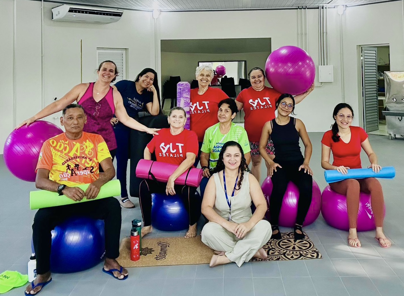 Imagem colorida mostra um grupo de dez pessoas em uma sala de ginástica. Elas estão sentadas ou de pé, cercadas por grandes bolas de Pilates e tapetes de ioga. Um homem está sentado à esquerda em uma bola com um tapete no colo. No centro, algumas mulheres estão de pé e uma está sentada entre elas. À direita, outras mulheres estão sentadas em bolas de Pilates, segurando tapetes de ioga. Outra mulher está sentada no chão entre os dois grupos. Todos parecem relaxados e felizes, posando para a foto em um ambiente de luz clara.