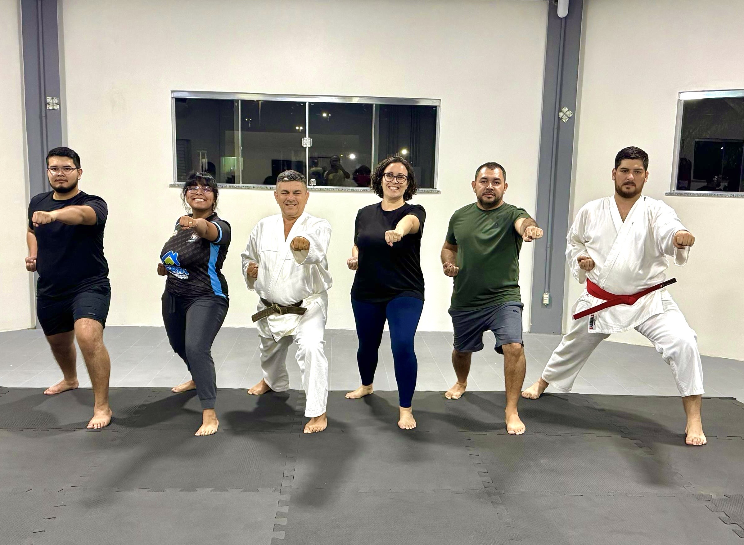Imagem colorida mostra seis pessoas em uma sala de ginástica. A maioria está descalça, em uma pose de caratê com o punho direito estendido para frente. Um homem está em uma posição de luta com a perna direita dobrada. Todos parecem concentrados.