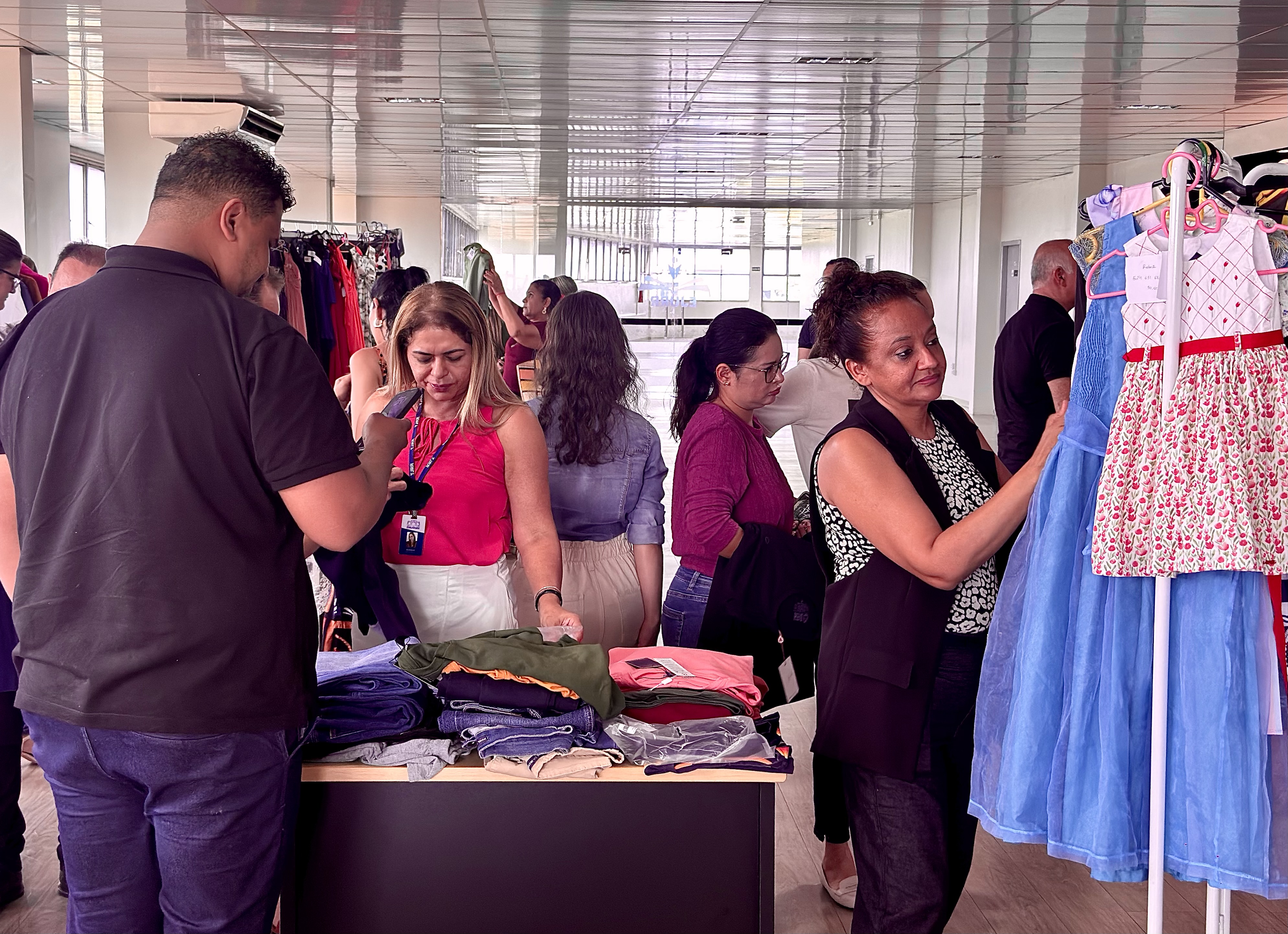 Imagem colorida mostra um grupo de pessoas em um espaço amplo e claro, onde está acontecendo um bazar de roupas. Há várias araras e mesas com pilhas de roupas. No primeiro plano, à esquerda, um homem de costas para a câmera mexe no celular. Ao centro, uma mulher de blusa rosa e calça branca olha para a mesa cheia de peças. No lado direito, uma mulher de blusa estampada, de perfil, mexe em uma arara de roupas. Ao fundo, outras pessoas circulam, observando as peças. O chão é de madeira clara e o teto é branco e metálico, com iluminação embutida.