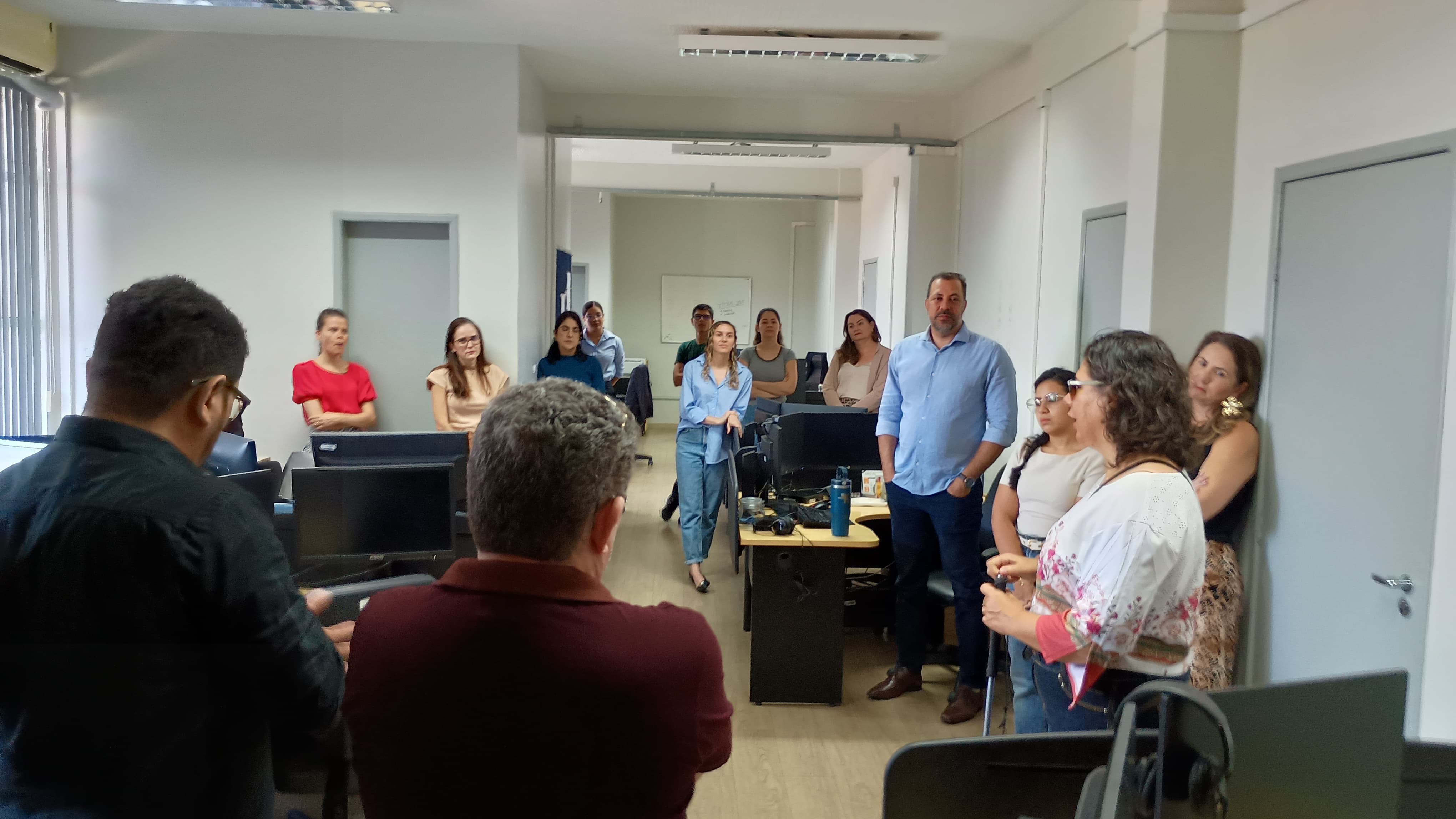 Imagem colorida mostra um escritório. Algumas pessoas estão sentadas em mesas com computadores, enquanto outras estão em pé, observando. O ambiente é um escritório de trabalho com divisórias e portas ao fundo.
