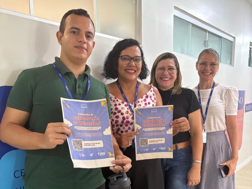 Foto colorida mostra Quatro pessoas estão em pé, lado a lado, sorrindo para a foto, em um corredor interno com piso de madeira clara e parede branca. À esquerda, um homem de camiseta polo verde-escura segura um cartaz da campanha de arrecadação de brinquedos. Ao lado dele, uma mulher de cabelo curto, óculos e blusa branca com estampa de bolinhas rosa também segura um cartaz da mesma campanha. Ambas as placas têm o título “Campanha de Arrecadação de Brinquedos” em destaque, com logotipos na parte inferior. As duas pessoas à direita sorriem e não seguram cartazes: uma mulher loira de saia cinza e blusa branca, e outra de cabelo loiro escuro, usando óculos e camiseta preta. Todas usam crachá azul pendurado no pescoço. Ao fundo, veem-se portas de vidro e janelas de divisórias de escritório.