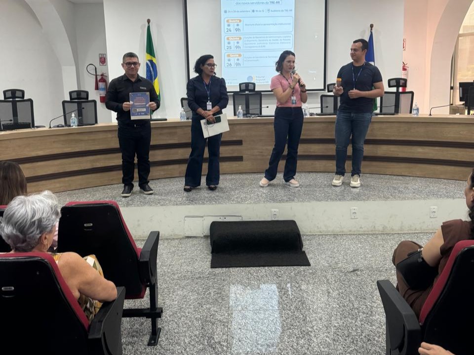 Foto colorida mostra em um auditório. Em pé, no palco, estão quatro servidores, dois homens e duas mulheres. Na frente do palco, pessoas assistem sentadas em cadeiras acolchoadas vermelhas. O piso é de granito claro e o ambiente está bem iluminado com luzes de teto.