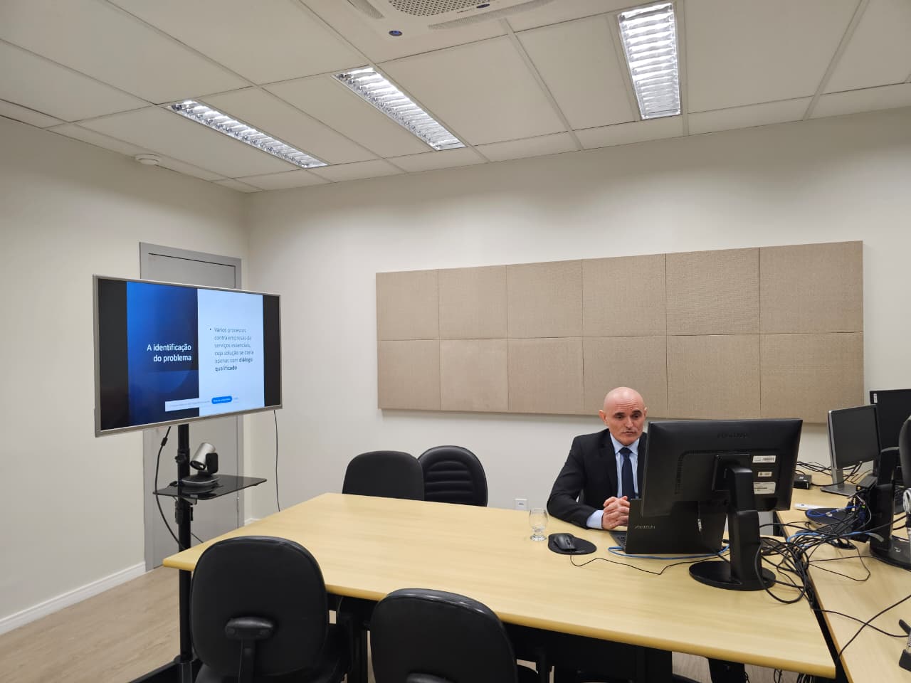 Imagem colorida mostra o juiz Air Marin sentado à mesa, com as mãos cruzadas e olhando para a tela à sua frente. Um televisor à sua esquerda exibe um slide com o título "A identificação do problema". A sala de reuniões é clara, com iluminação de teto, painéis acústicos na parede e microfones sobre a mesa.