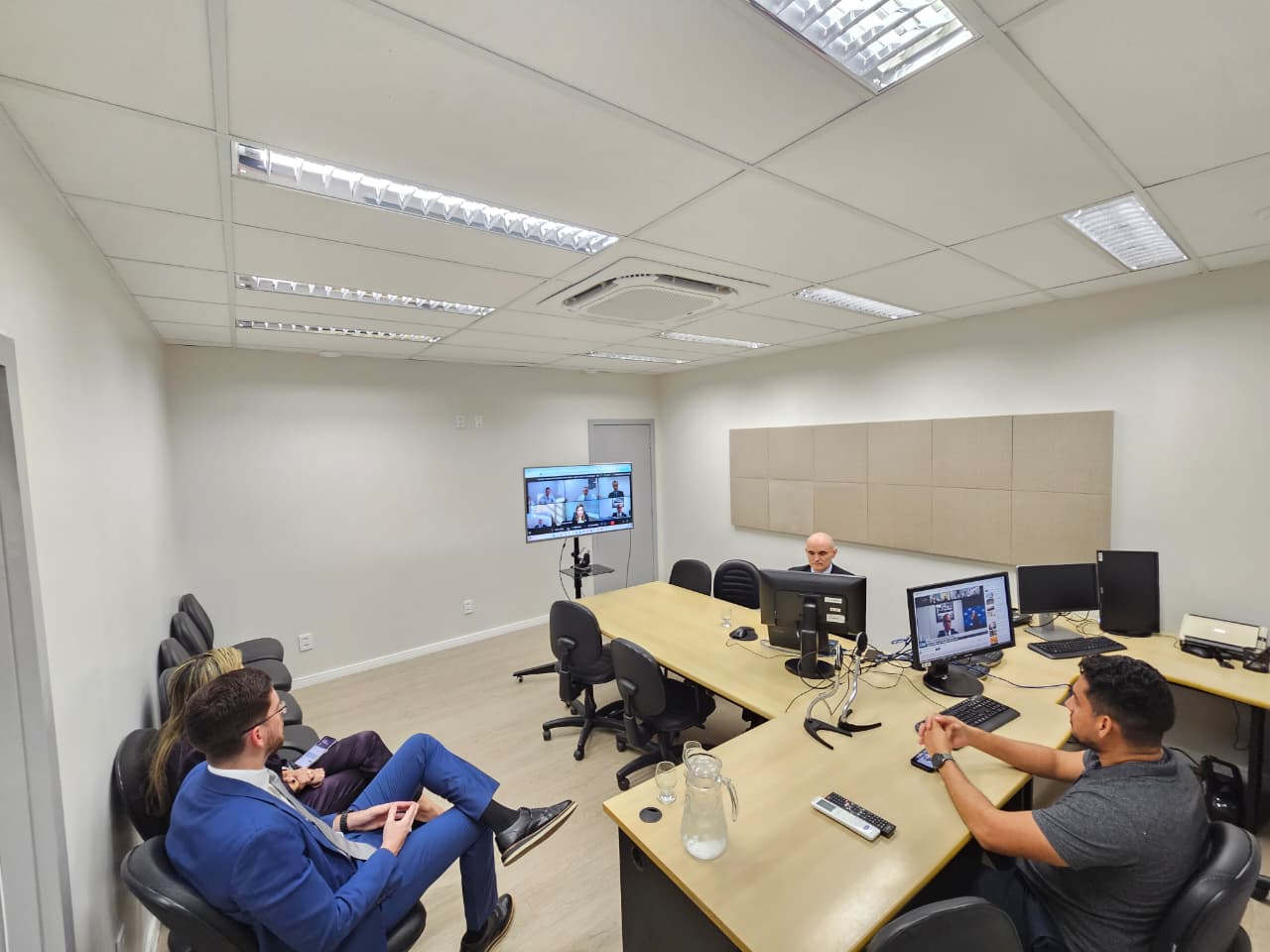 Imagem colorida de uma sala de reunião com paredes claras e teto branco. Pessoas estão sentadas em torno de uma mesa com computadores. Em primeiro plano, um homem de terno azul está sentado, e outro homem, de camisa cinza, olha para um monitor. Ao fundo, uma tela maior em um pedestal exibe uma videoconferência.