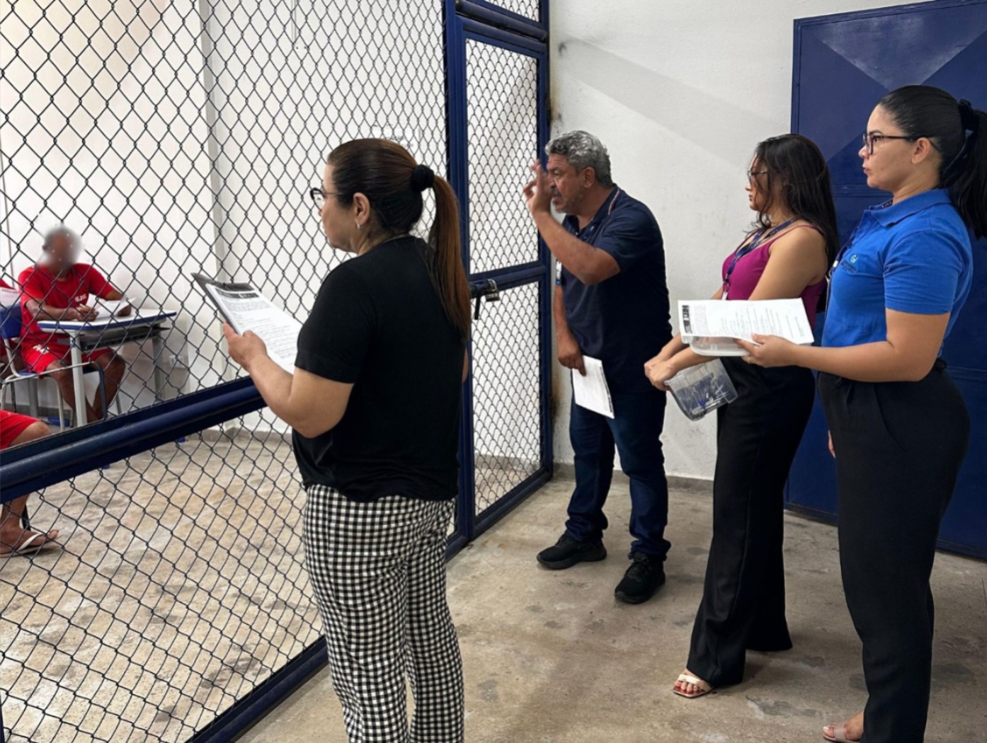 A imagem mostra quatro pessoas posicionadas em frente a uma grade de metal pintada de azul, sendo um homem e três mulheres. Uma das mulheres, de cabelos presos e óculos, segura um questionário; ao seu lado, um homem de barba grisalha faz um gesto com a mão, como se estivesse explicando ou dando orientações. Duas outras mulheres, uma com blusa roxa e outra com camisa azul, também seguram documentos. Do outro lado da grade, presos uniformizados em vermelho estão sentados em carteiras escolares individuais.