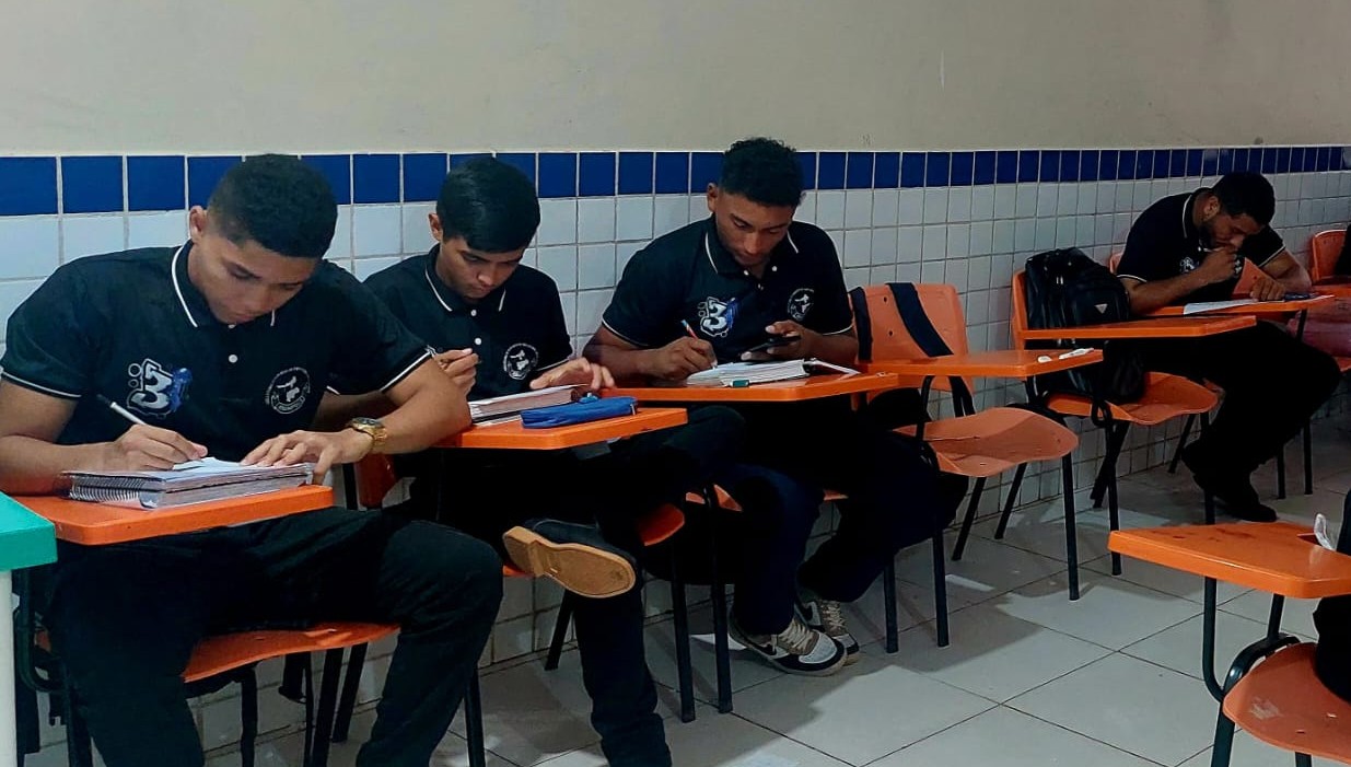  imagem mostra quatro estudantes em uma sala de aula. Eles vestem uniformes pretos com golas e detalhes brancos, que trazem estampados o logotipo da instituição. Sentados em carteiras escolares individuais de cor laranja, respondem questionários aplicados pela pesquisa.