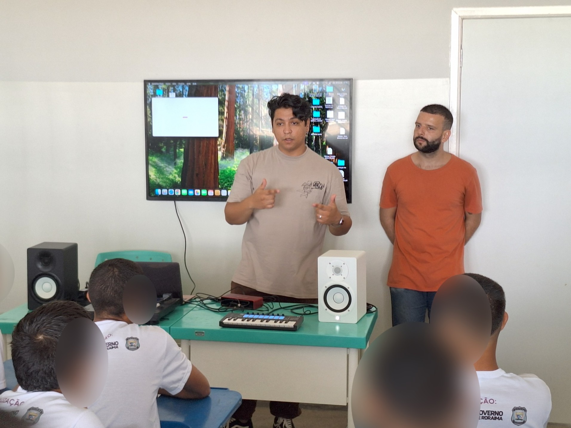 Foto colorida mostra homens adultos à frente de uma sala de aula, em pé diante de uma mesa com equipamentos de produção musical, como teclado, caixas de som e notebook. Um deles, de camiseta bege, fala gesticulando com as mãos, enquanto o outro, de camiseta laranja, observa. Ao fundo, há uma televisão com a imagem de uma floresta exibida como plano de fundo. Sentados, vários jovens com camisetas brancas d. Os rostos dos adolescentes estão desfocados para preservar a identidade. Foto colorida mostra homens adultos à frente de uma sala de aula, em pé diante de uma mesa com equipamentos de produção musical, como teclado, caixas de som e notebook. Um deles, de camiseta bege, fala gesticulando com as mãos, enquanto o outro, de camiseta laranja, observa. Ao fundo, há uma televisão com a imagem de uma floresta exibida como plano de fundo. Sentados, vários jovens com camisetas brancas d. Os rostos dos adolescentes estão desfocados para preservar a identidade.