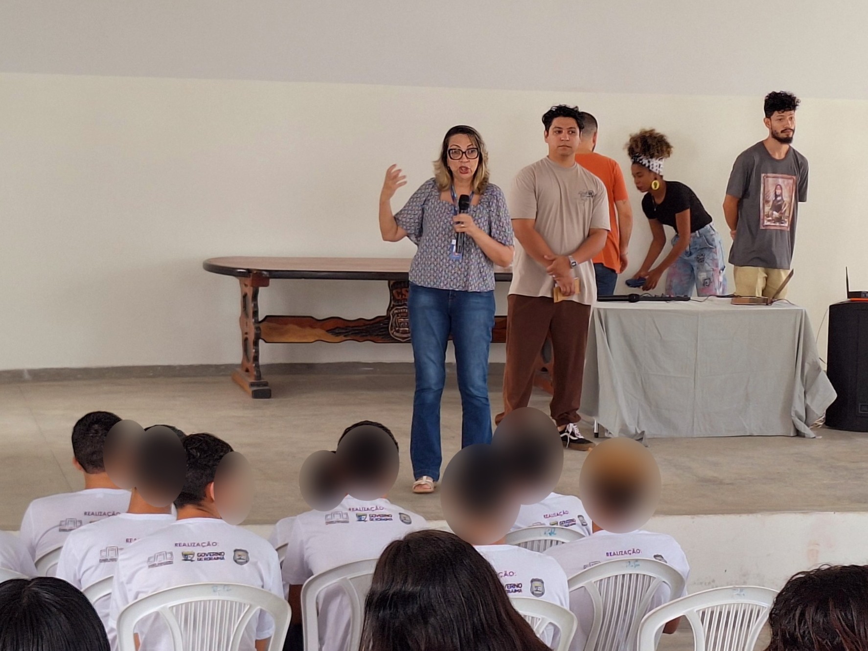 Foto colorida mostra uma mulher de óculos, vestindo blusa estampada azul e calça jeans, falando ao microfone em um palco de auditório. Atrás dela, há uma mesa com toalha e equipamentos, onde quatro jovens (dois homens e duas mulheres) se preparam para atividades culturais. Um dos rapazes veste camiseta com estampa de arte religiosa. Ao fundo, adolescentes com camisetas brancas do sistema socioeducativo assistem sentados em cadeiras plásticas brancas. Os rostos dos adolescentes foram desfocados para preservar suas identidades. Foto colorida mostra uma mulher de óculos, vestindo blusa estampada azul e calça jeans, falando ao microfone em um palco de auditório. Atrás dela, há uma mesa com toalha e equipamentos, onde quatro jovens (dois homens e duas mulheres) se preparam para atividades culturais. Um dos rapazes veste camiseta com estampa de arte religiosa. Ao fundo, adolescentes com camisetas brancas do sistema socioeducativo assistem sentados em cadeiras plásticas brancas. Os rostos dos adolescentes foram desfocados para preservar suas identidades.