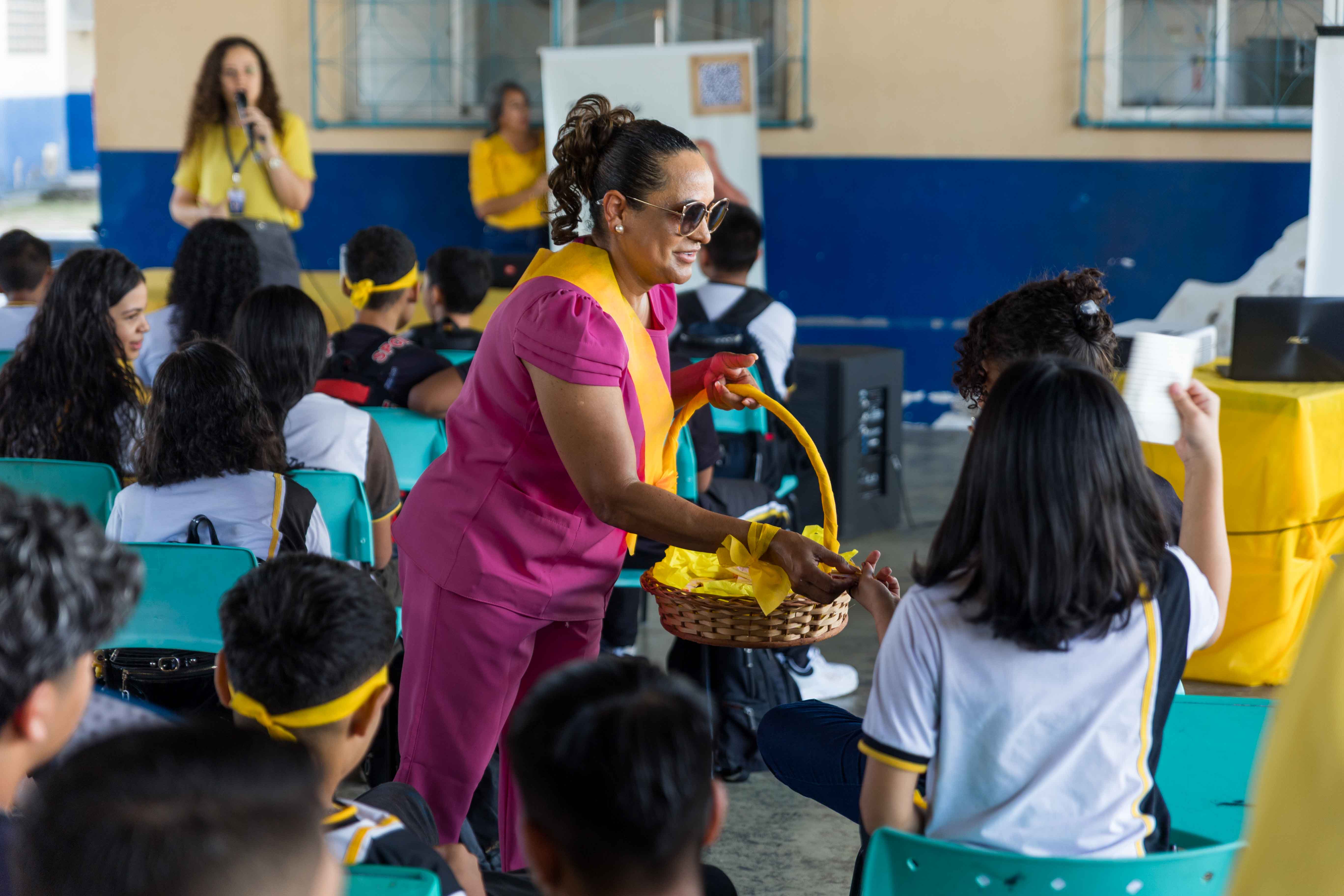  Imagem colorida mostra uma mulher de óculos escuros e uniforme rosa e amarelo, segurando uma cesta de vime com fitas amarelas. Ela se inclina para oferecer algo a uma jovem, enquanto outros estudantes assistem. A cena acontece em um ambiente escolar, com uma mulher ao fundo falando em um microfone.
