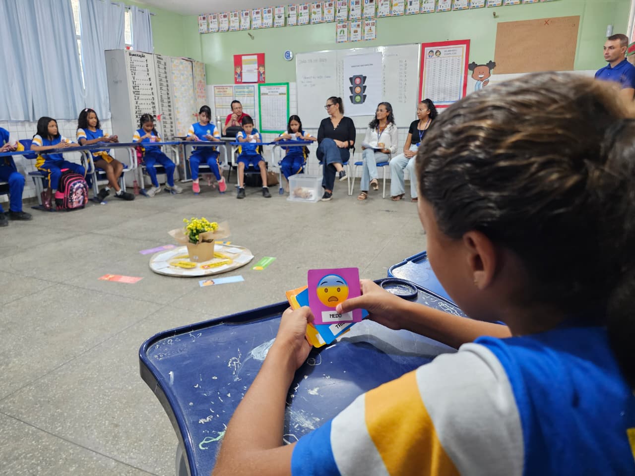  Foto colorida mostra o momento de uma roda de conversa em sala de aula, com foco em uma aluna que segura cartas coloridas com carinhas de emoções. Em destaque, uma carta roxa com o emoji de expressão assustada e a palavra “MEDO”. Ao fundo, crianças e adultos participam da atividade, sentados em círculo. No centro, há um arranjo com flores e cartões coloridos no chão.
