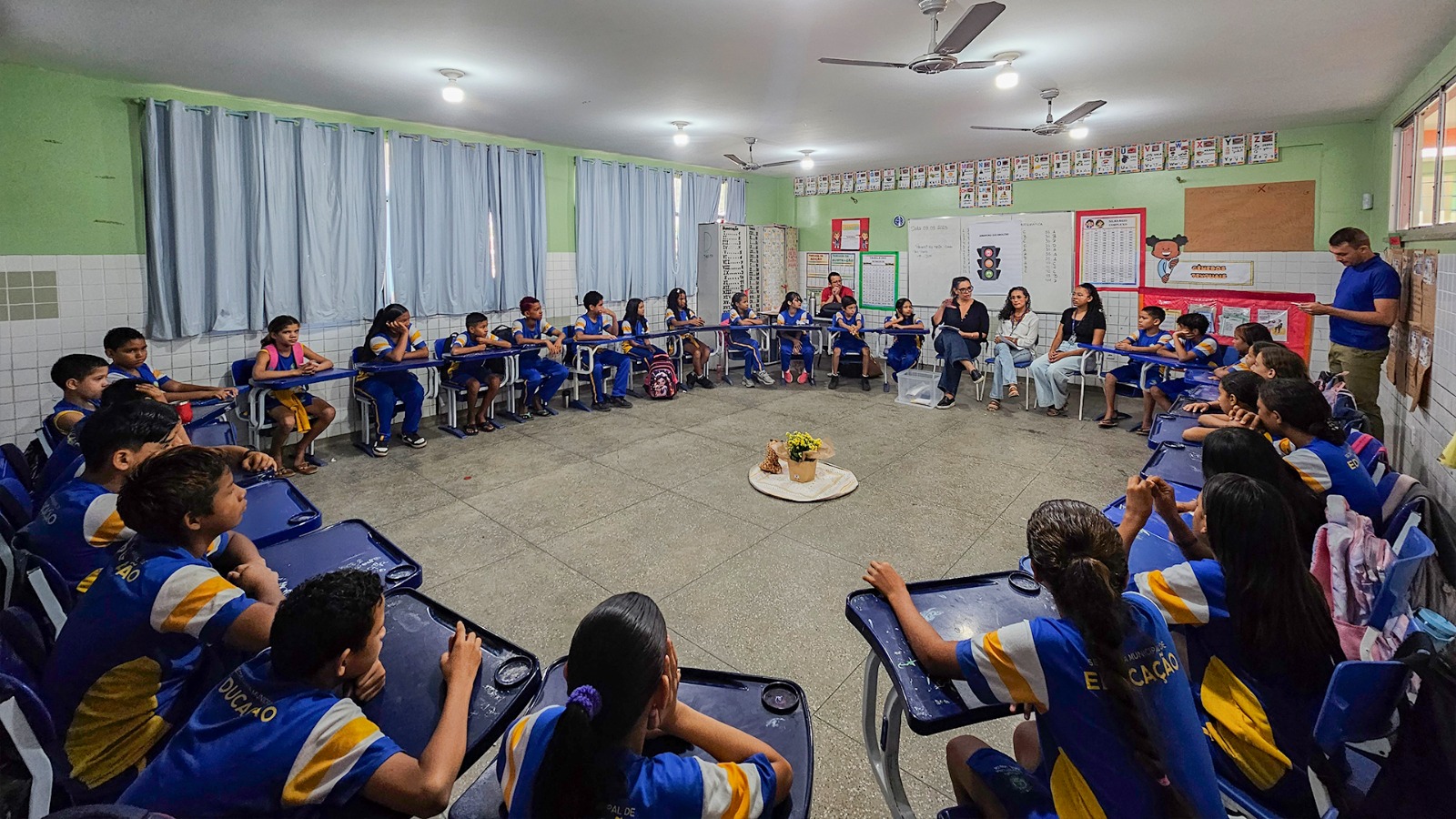 Foto colorida mostra uma roda de conversa em sala de aula com cerca de 29 crianças uniformizadas e cinco adultos. Todos estão sentados em cadeiras dispostas em círculo, num ambiente acolhedor. No centro da roda, há uma pequena decoração com flores amarelas e tecido claro. A sala tem paredes verdes, cartazes educativos e ventiladores no teto.