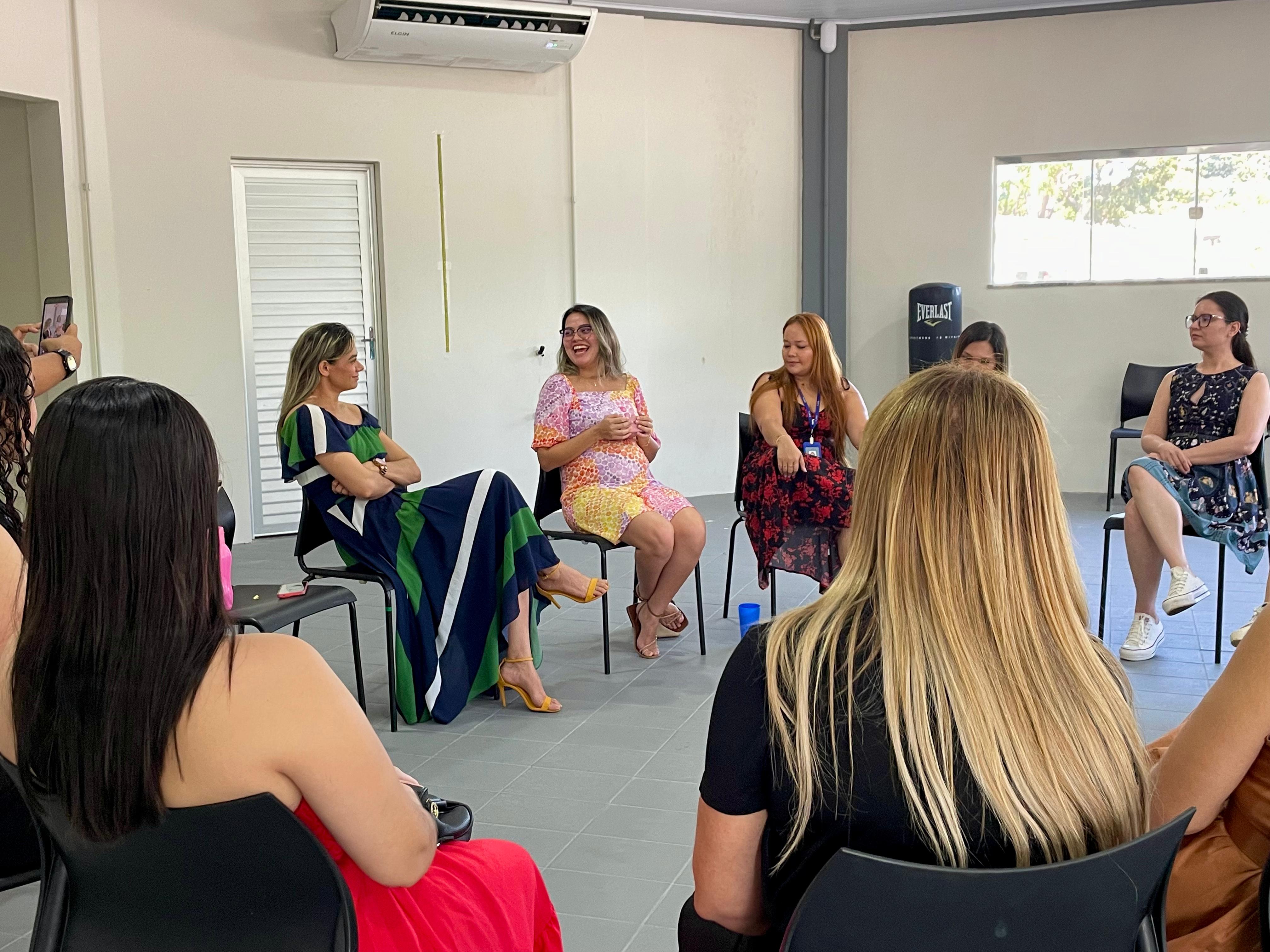 A imagem mostra um grupo de mulheres reunidas em círculo dentro de uma sala clara, de paredes brancas e piso cinza. Todas estão sentadas em cadeiras de plástico pretas, posicionadas de forma a favorecer a conversa. Em primeiro plano, vemos mulheres de costas, uma delas com cabelo loiro comprido e outra de vestido vermelho. No centro da imagem, à esquerda, uma delas veste um vestido longo listrado em azul, verde e branco, ao lado, outra mulher sorri enquanto fala, usando um vestido curto estampado em rosa, amarelo e laranja, com sandálias amarelas. À direita, há outras participantes, incluindo uma mulher de vestido preto com flores vermelhas e outra de vestido azul estampado com tênis brancos. No canto esquerdo, alguém segura um celular, registrando o momento. Ao fundo, há uma janela que deixa entrar a luz natural e um aparelho de ar-condicionado fixado na parede.