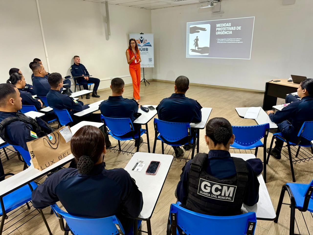 Imagem colorida mostra um grupo de pessoas em uniformes azuis, com a sigla GCM (Guarda Civil Municipal) nas costas, sentadas em carteiras azuis em uma sala de aula. Elas estão atentas a uma palestrante, que está de pé, vestindo uma calça e um blazer laranja. Atrás dela, um projetor exibe uma apresentação com o título "MEDIDAS PROTETIVAS DE URGÊNCIA". A cena retrata um momento de treinamento ou capacitação profissional. Imagem colorida mostra um grupo de pessoas em uniformes azuis, com a sigla GCM (Guarda Civil Municipal) nas costas, sentadas em carteiras azuis em uma sala de aula. Elas estão atentas a uma palestrante, que está de pé, vestindo uma calça e um blazer laranja. Atrás dela, um projetor exibe uma apresentação com o título "MEDIDAS PROTETIVAS DE URGÊNCIA". A cena retrata um momento de treinamento ou capacitação profissional.