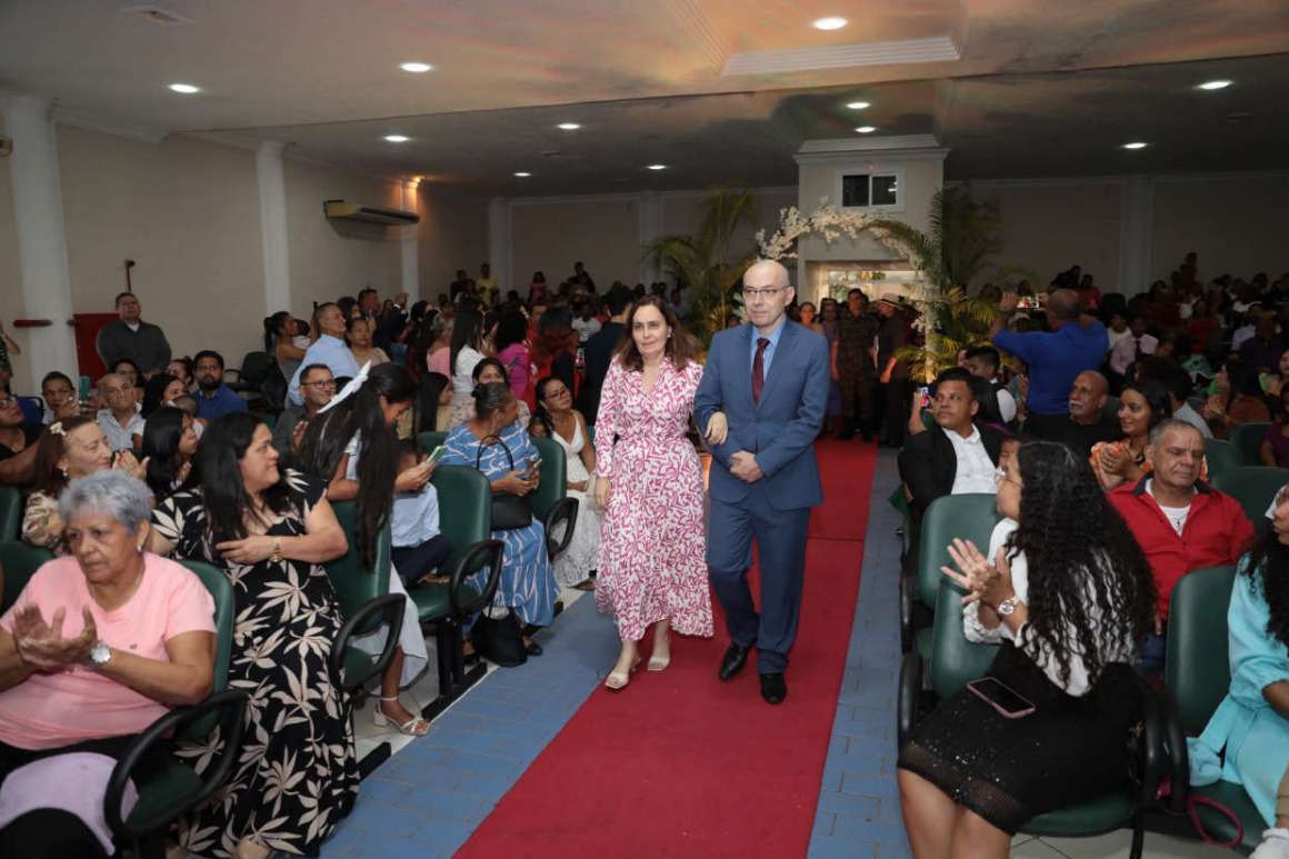 Foto colorida mostra o desembargador Erick Linhares, corregedor-geral de justiça do TJRR, caminhando de braços dados com sua esposa, por um corredor com tapete vermelho em um auditório lotado.    