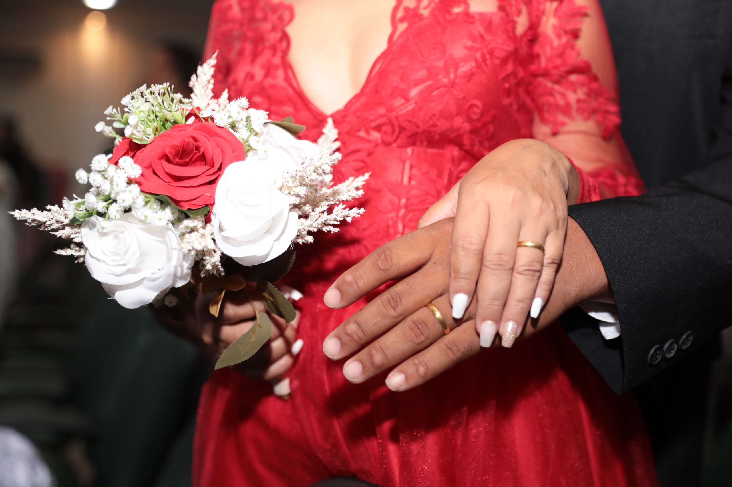 Foto colorida mostra em primeiro plano, um casal de mãos dadas, com as alianças de casamento em destaque. A noiva, com vestido de renda vermelho e unhas pintadas de branco, segura um buquê com rosas vermelhas e brancas. O noivo veste terno preto.