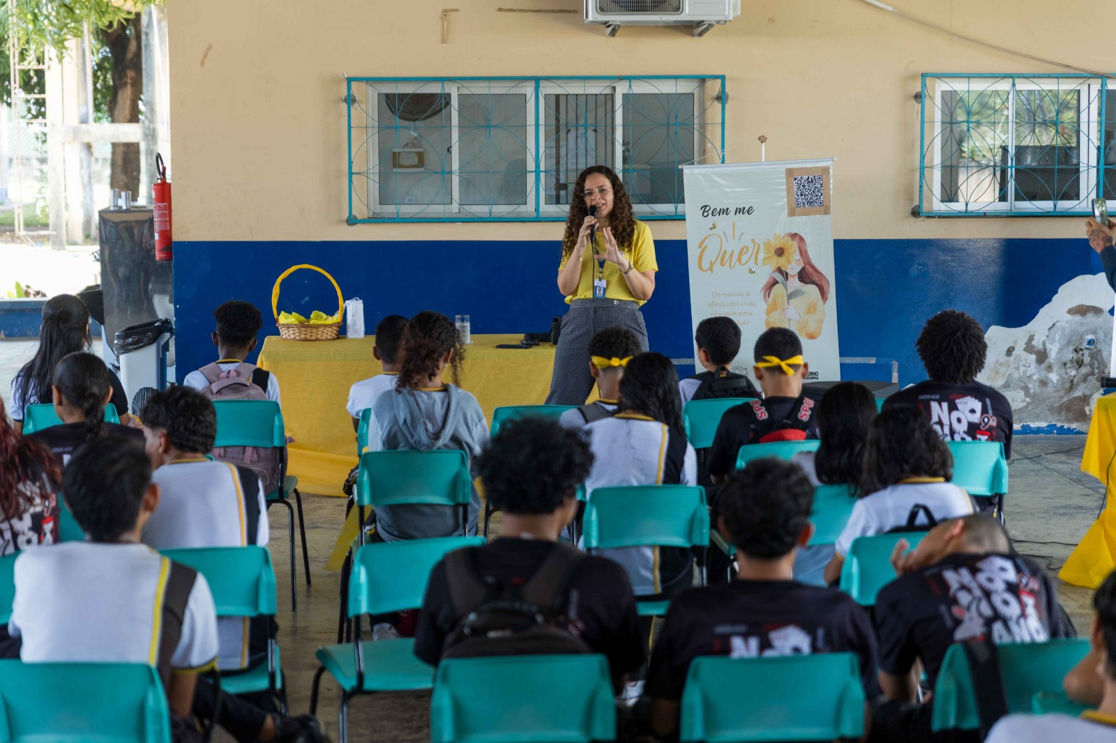 A imagem colorida de uma mulher está em pé, falando ao microfone, para uma plateia de jovens (estudantes) sentados em cadeiras de plástico verde-água. Atrás dela, há um banner com a frase "Bem me Quer" e um código QR. O evento acontece em uma área coberta com paredes de cores bege e azul, sugerindo ser uma atividade educacional ou informativa para adolescentes.