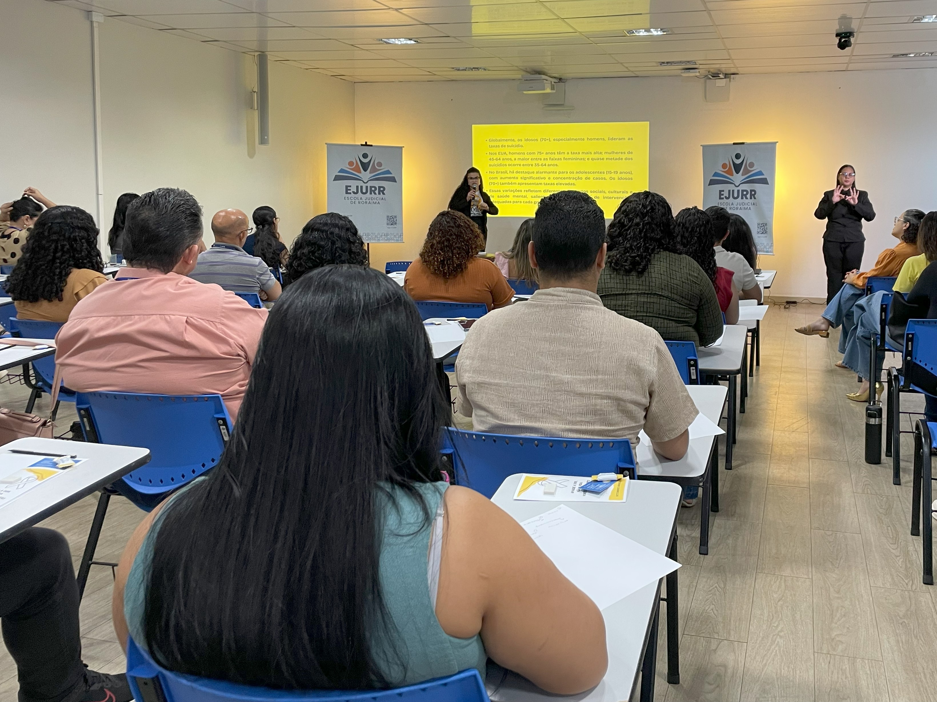 A imagem colorida mostra uma sala, com participantes sentados em cadeiras escolares azuis, voltados para o fundo da sala, onde ocorre uma palestra. No centro do ambiente, uma mulher fala ao microfone, enquanto há uma do outro lado fazendo libras. 