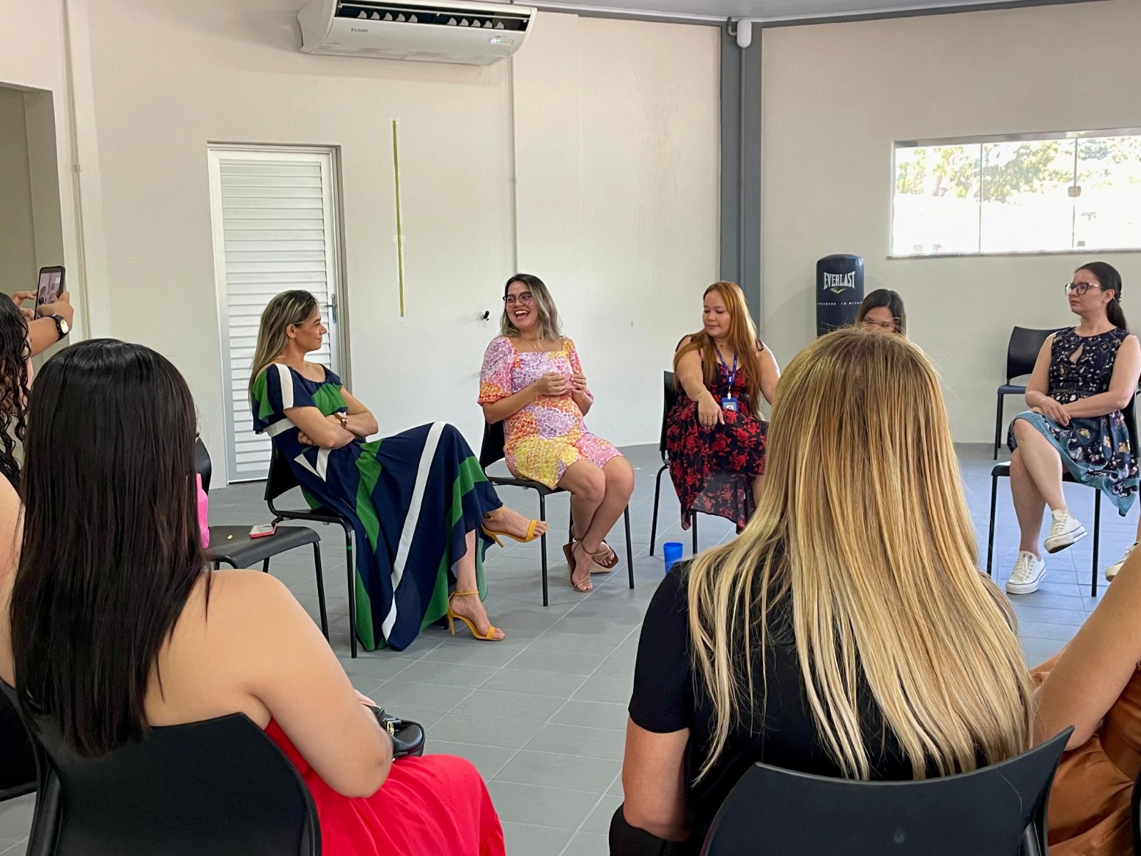 A imagem mostra um grupo de mulheres reunidas em círculo dentro de uma sala clara, de paredes brancas e piso cinza. Todas estão sentadas em cadeiras de plástico pretas, posicionadas de forma a favorecer a conversa.