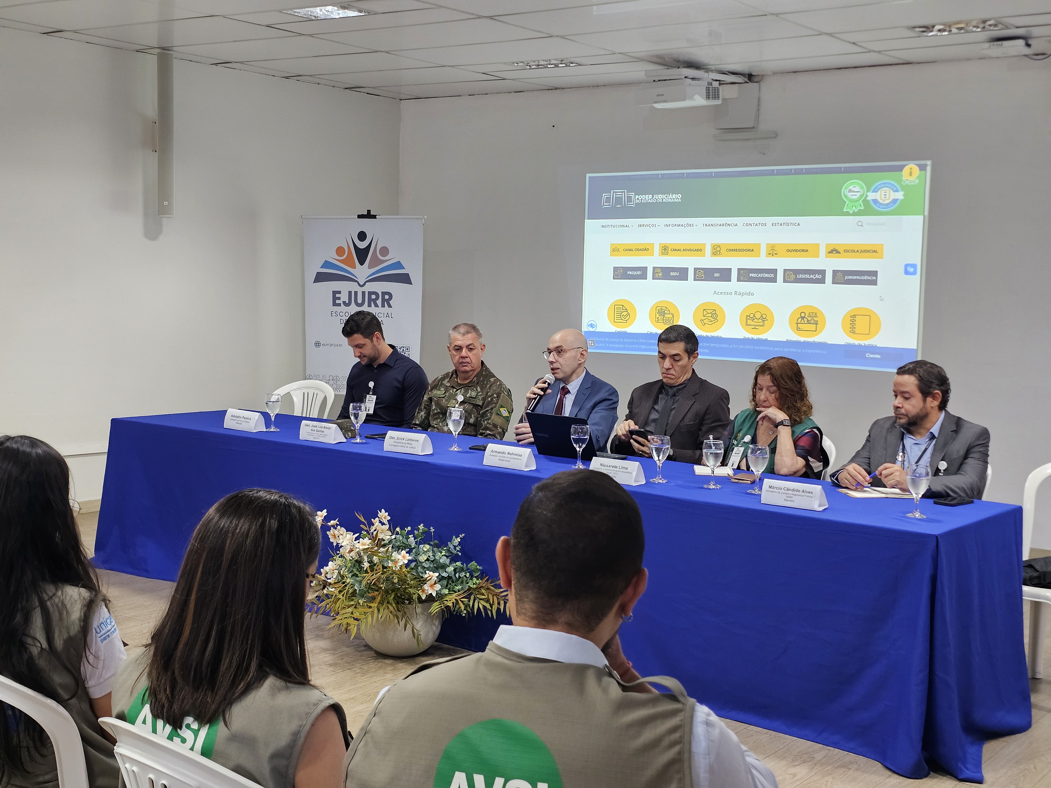 Foto colorida mostra uma mesa solene composta por seis pessoas sentadas, participando de um evento institucional em uma sala da Escola do Poder Judiciário de Roraima. Foto colorida mostra uma mesa solene composta por seis pessoas sentadas, participando de um evento institucional em uma sala da Escola do Poder Judiciário de Roraima.