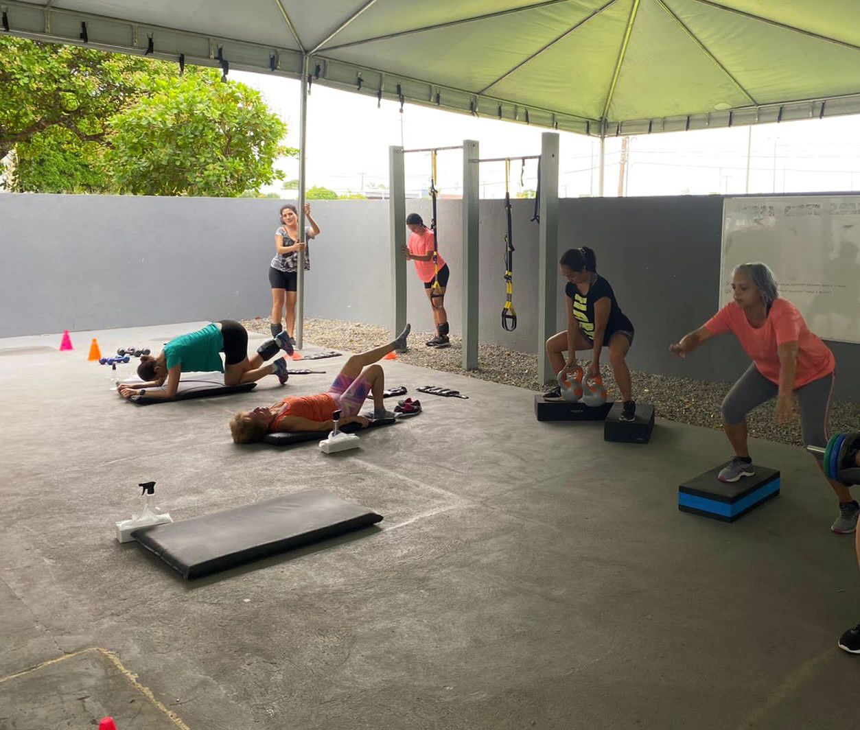 Imagem colorida mostra um grupo de seis mulheres praticando exercícios em uma academia funcional ao ar livre. No primeiro plano, uma mulher se prepara para um agachamento enquanto outras realizam prancha, abdominais e levantamento de kettlebell. O treino acontece sob um toldo em um ambiente com piso de cimento e um muro cinza ao fundo.