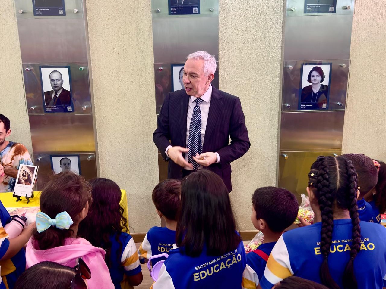 A imagem mostra um hall interno iluminado, onde o vice-presidente do TJRR, desembargador Almiro Padilha, conversa com um grupo de crianças. Ele está de frente para os alunos, gesticulando com as mãos enquanto fala. As crianças, vestidas com uniformes azuis e amarelos escutam atentamente. Ao fundo, há uma parede com retratos emoldurados de ex-gestores e autoridades do Tribunal de Justiça de Roraima.