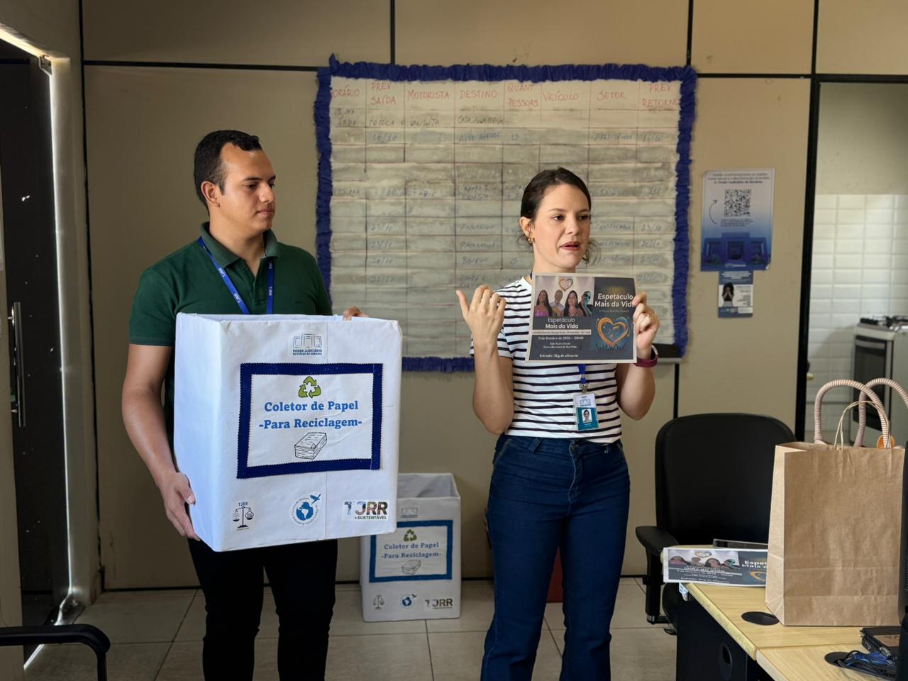 Imagem colorida mostra um homem e uma mulher, ambos em pé no centro de uma sala. O homem segura uma caixa de papelão branca com os dizeres “coletor de papel para reciclagem”, com a inscrição em azul. A mulher ao seu lado segura um folder ou cartaz colorido com o título "Expectativa - Mais de Vida" e fotos de pessoas. Atrás deles, na parede, há um grande quadro de avisos com papéis e anotações manuscritas em uma grade. Um segundo coletor de papel menor está no chão, à direita da mulher.