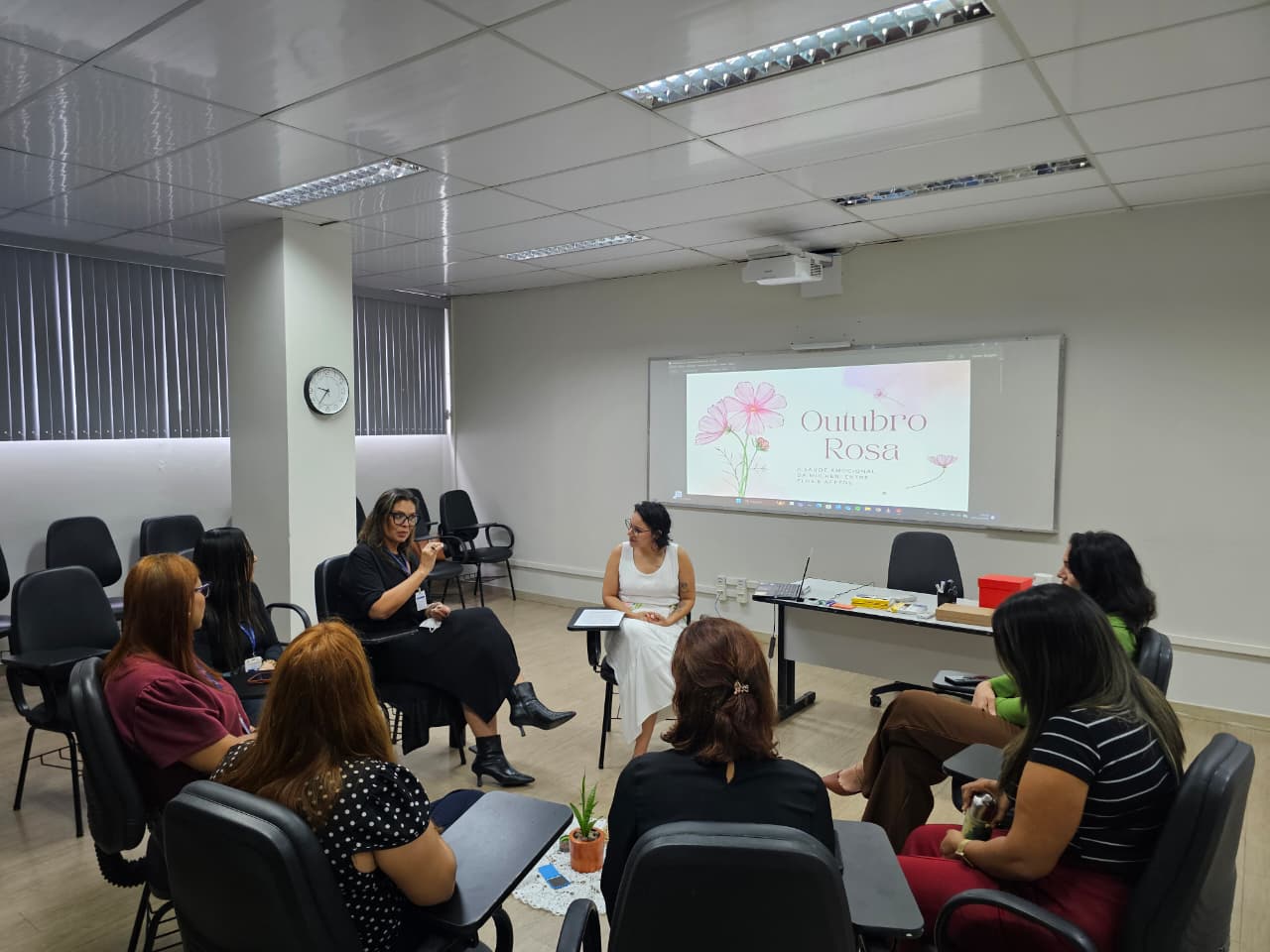 A imagem colorida mostra uma roda de conversa com nove mulheres, reunidas em uma sala de reuniões clara e organizada. As cadeiras estão dispostas em círculo, promovendo um ambiente de diálogo acolhedor. No centro, uma mulher de vestido branco comanda a conversa.