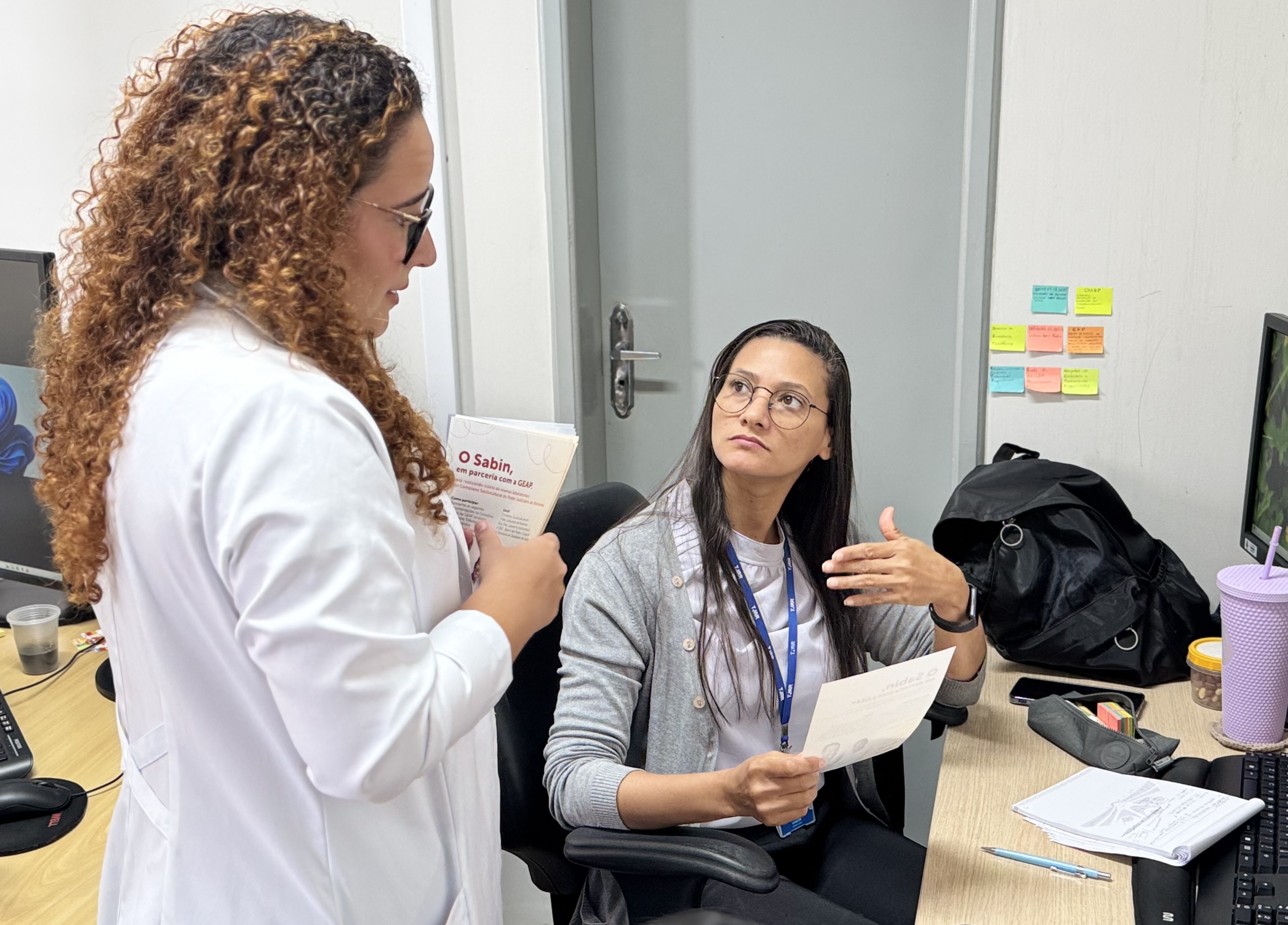 Foto colorida mostra à esquerda, uma profissional de jaleco branco e cabelos cacheados segurando panfletos informativos do Sabin e da GEAP está de pé, dialogando com a outra mulher, que está sentada em frente a uma mesa. A mulher sentada usa óculos, crachá azul e veste camiseta branca com cardigã cinza claro. Ela segura um dos panfletos, escutando com atenção, enquanto gesticula com a mão livre.