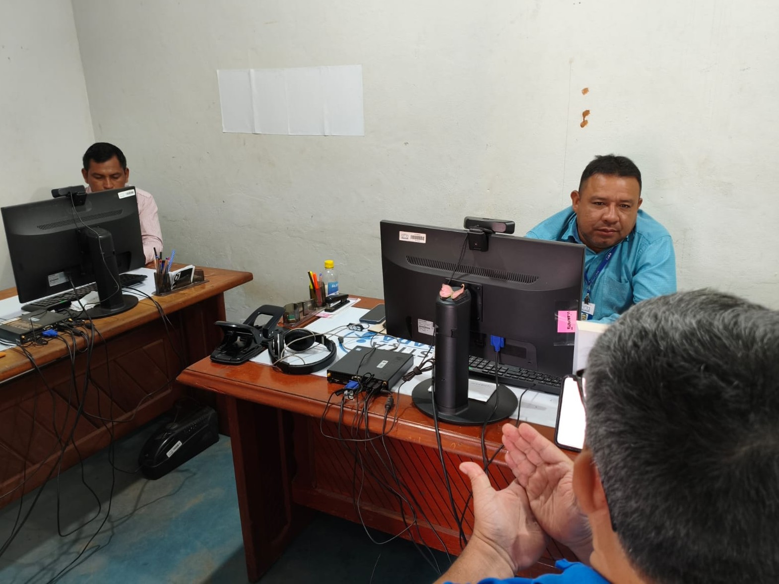 imagem colorida em ambiente interno de uma reunião entre três servidores do TJRR.  Dois deles estão sentados em frente a computadores e outro está de costas, participando da conversa. Sobre as mesas há diversos equipamentos e materiais de trabalho. 
