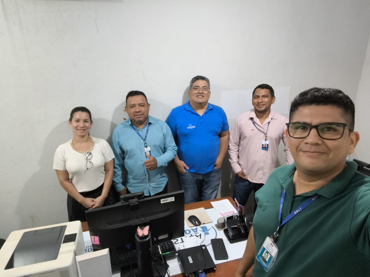 Em um escritório, dois homens estão sentados diante de computadores e outro, de costas, conversa com eles. As mesas têm cabos, papéis e materiais de trabalho.