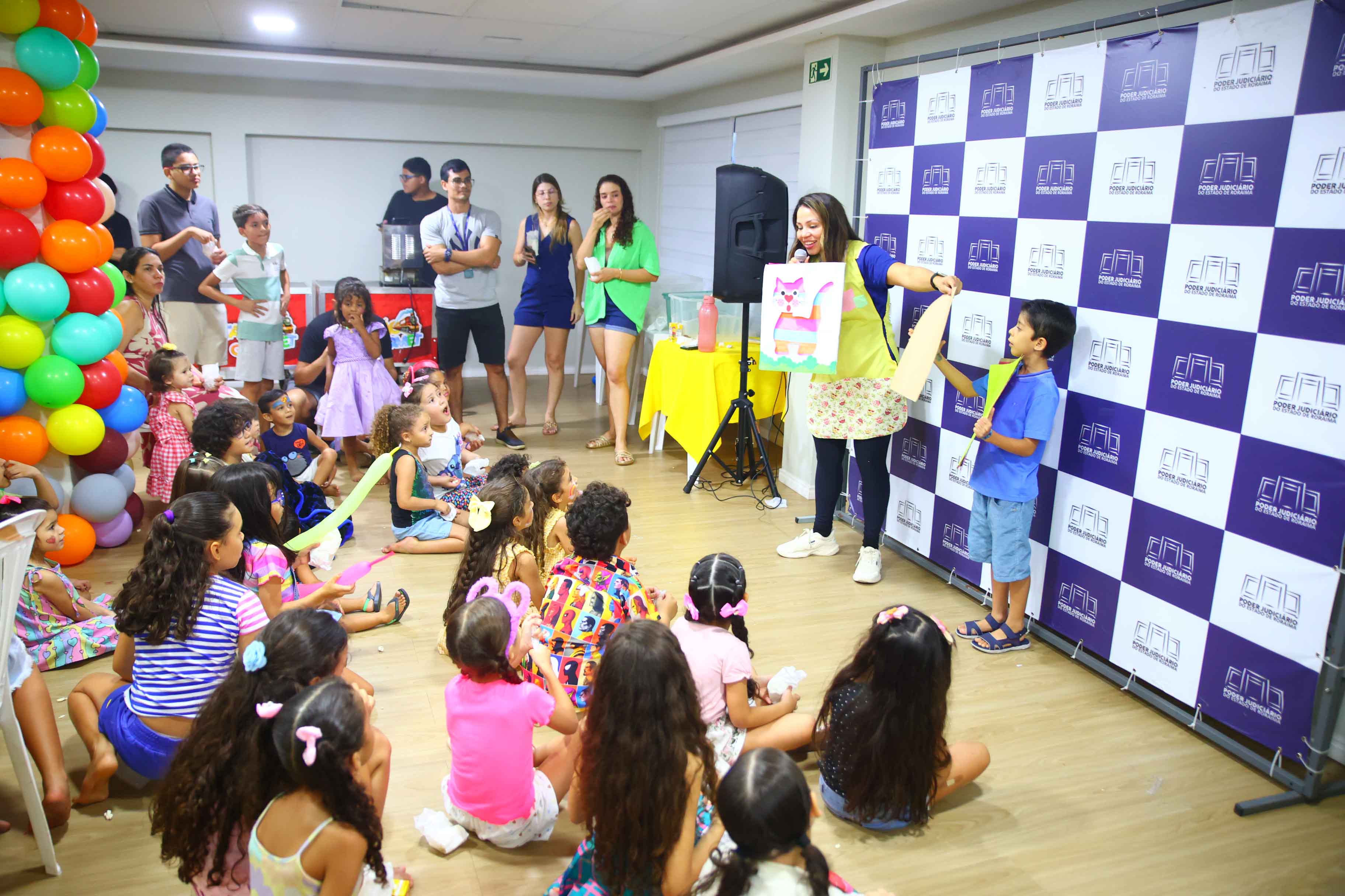 Imagem colorida das um grupo de crianças sentadas no chão ouvindo a contação de história. A frente há uma servidora da SQV contando uma história segurando o cartaz de um gato
