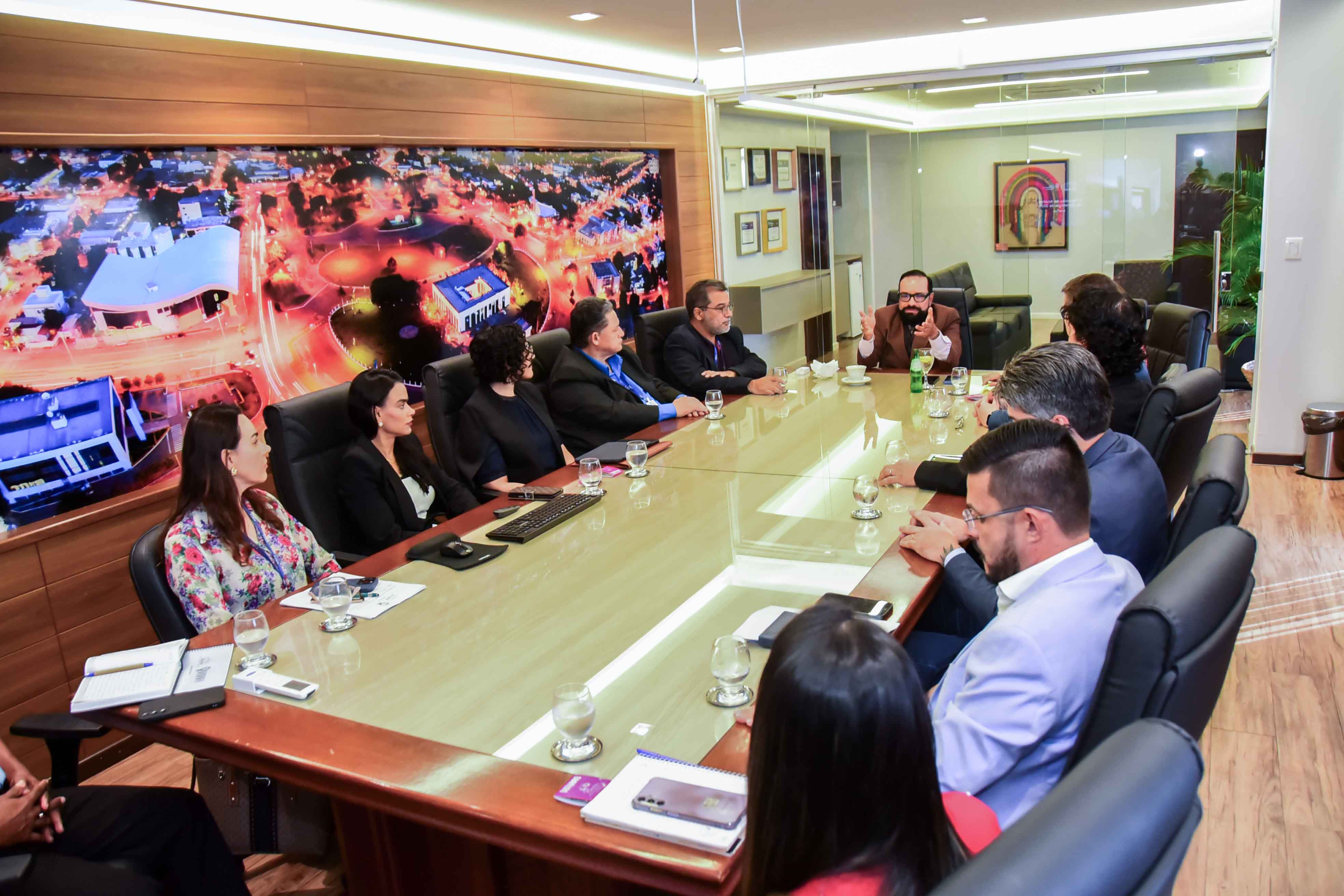  Imagem mostra uma reunião em uma sala ampla e bem iluminada, com uma grande mesa de madeira e tampo de vidro. Cerca de doze pessoas estão sentadas ao redor. No centro da mesa, há copos com água, notebooks, anotações e um teclado. As pessoas vestem trajes formais, como ternos, blazers e camisas sociais. Ao fundo, há uma parede de vidro com persianas verticais e uma planta decorativa. O ambiente tem um ar institucional e profissional, sugerindo uma reunião de trabalho ou encontro entre gestores.