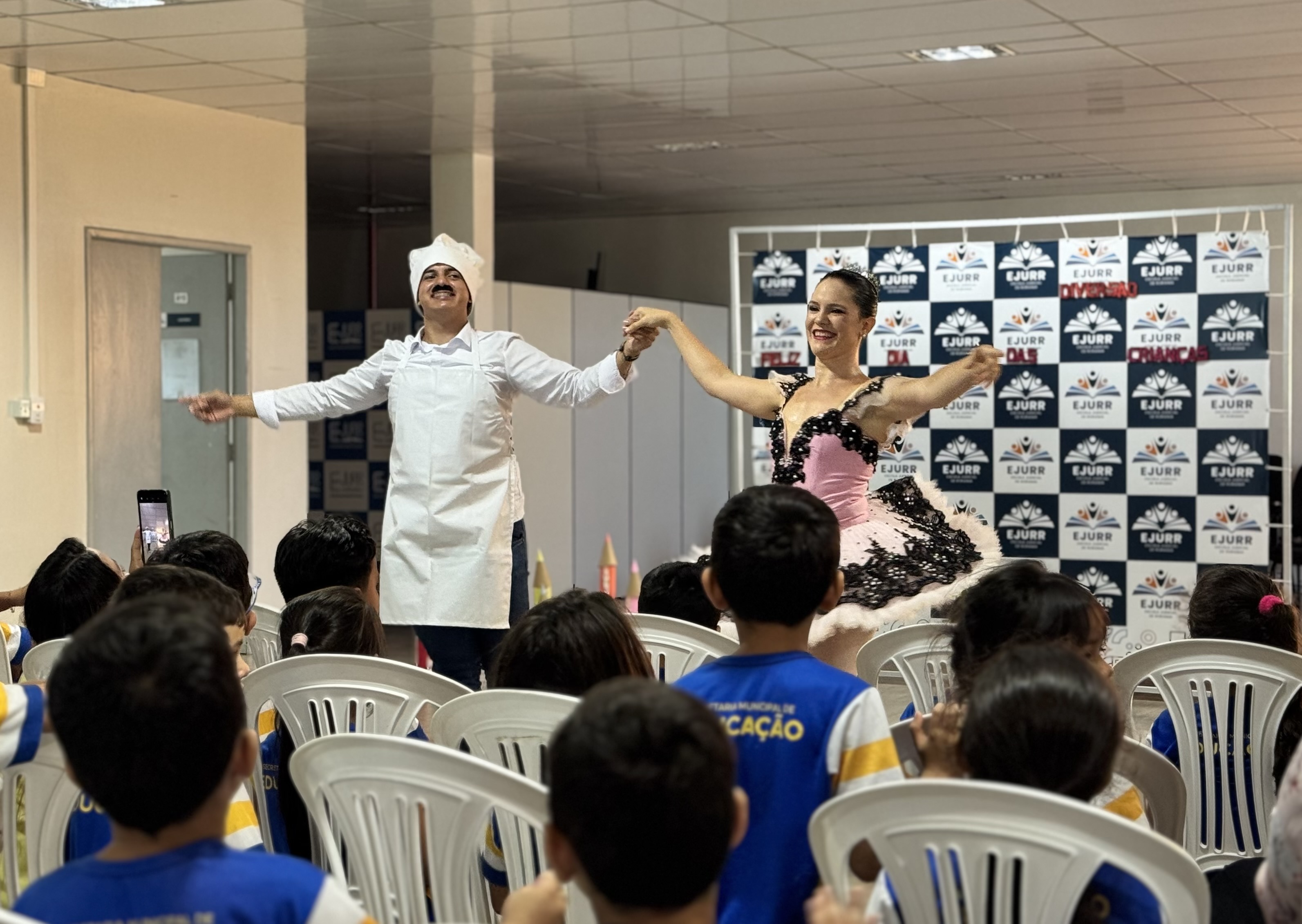 Crianças assistem a uma apresentação de dança ou teatro, onde um homem vestido de cozinheiro e uma mulher com vestido de festa se apresentam em frente a um painel temático do TJRR/EJURR. Crianças assistem a uma apresentação de dança ou teatro, onde um homem vestido de cozinheiro e uma mulher com vestido de festa se apresentam em frente a um painel temático do TJRR/EJURR.