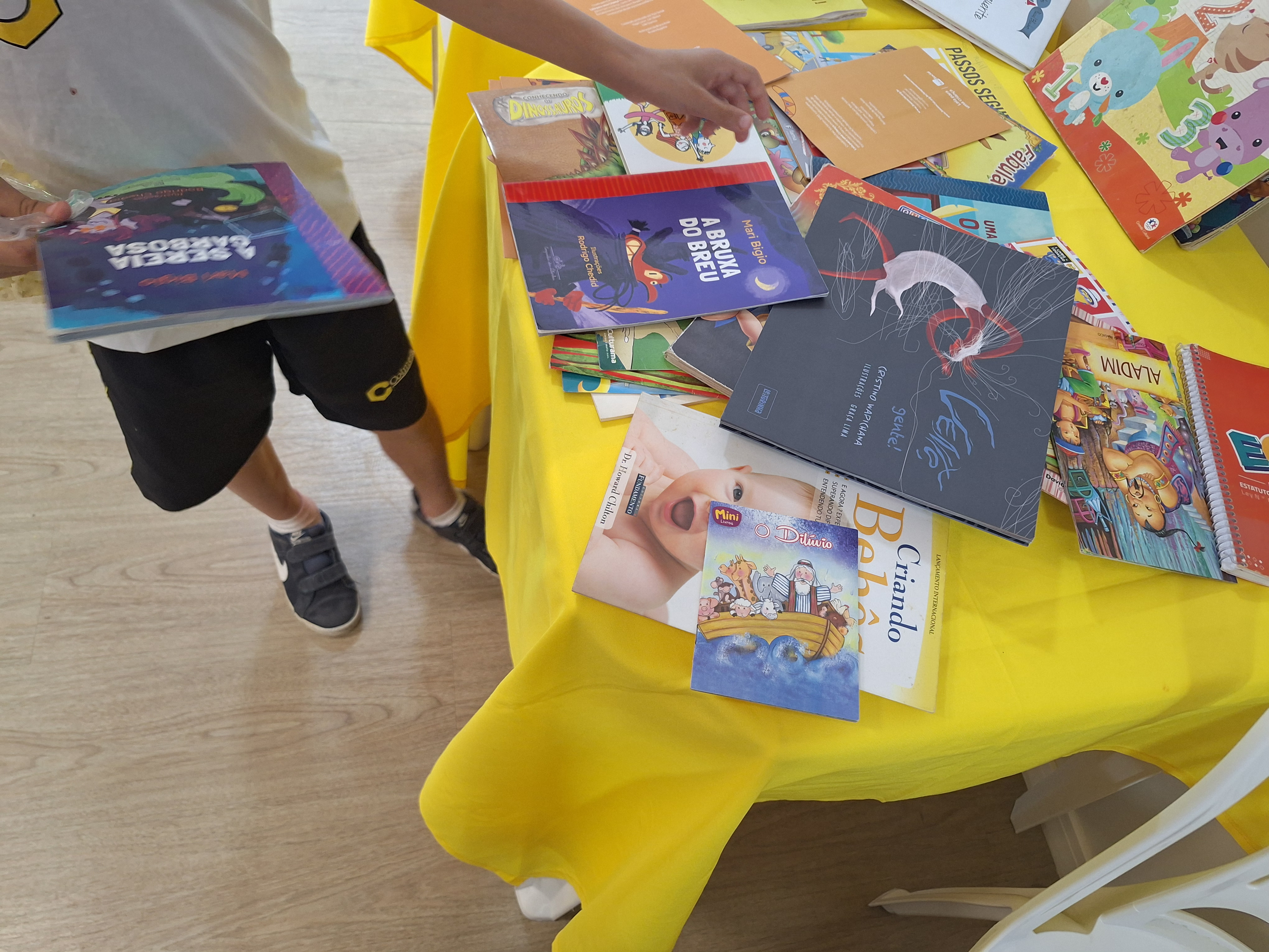   A imagem colorida mostra uma mesa coberta com toalha amarela, onde vários livros infantis estão espalhados. As capas coloridas exibem ilustrações de personagens, animais. À esquerda, uma criança em pé, vestindo camiseta branca e shorts escuros, folheia um dos livros. O ambiente é claro e transmite um clima de curiosidade e incentivo à leitura infantil.   A imagem colorida mostra uma mesa coberta com toalha amarela, onde vários livros infantis estão espalhados. As capas coloridas exibem ilustrações de personagens, animais. À esquerda, uma criança em pé, vestindo camiseta branca e shorts escuros, folheia um dos livros. O ambiente é claro e transmite um clima de curiosidade e incentivo à leitura infantil. 