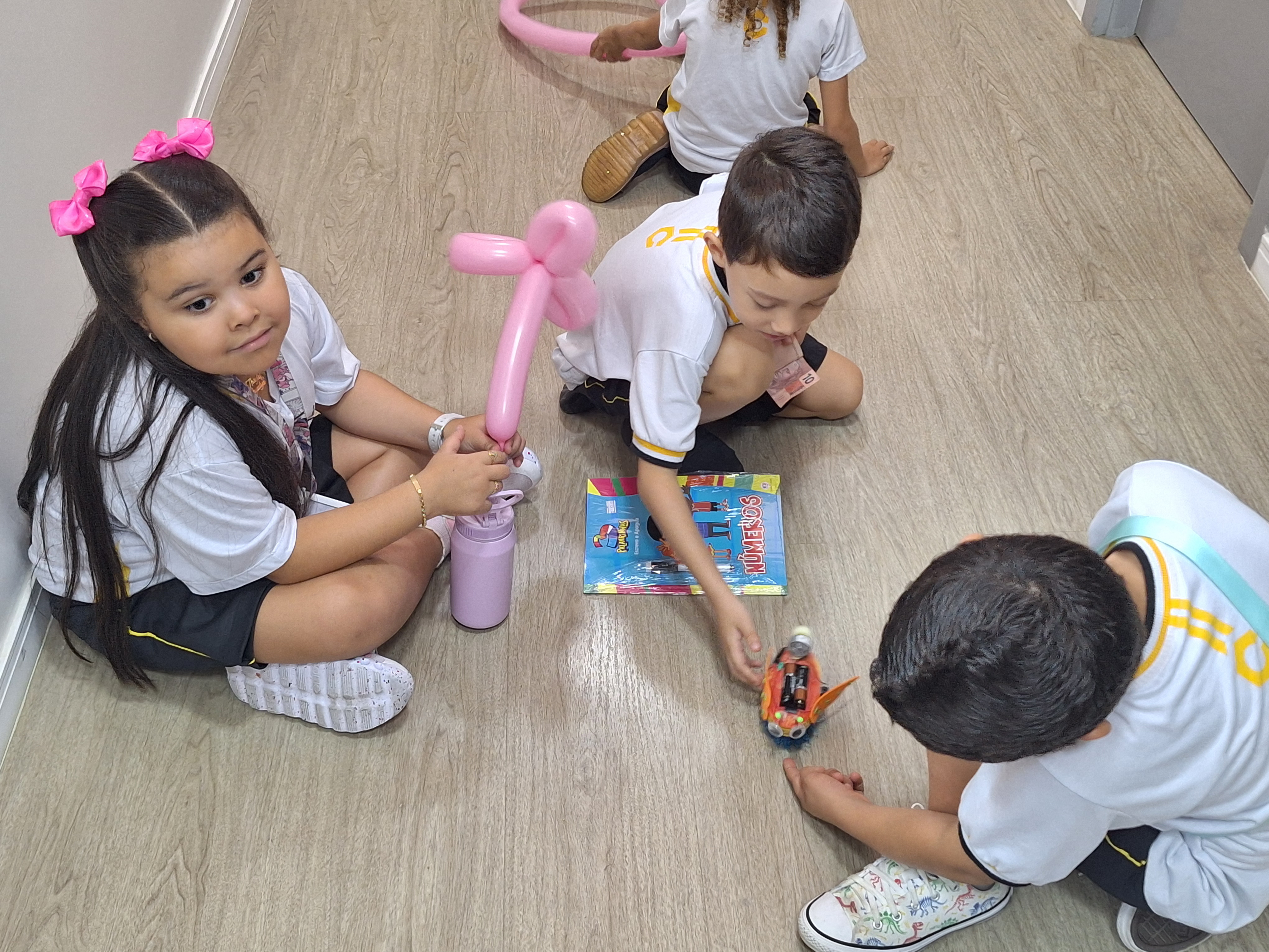 A imagem colorida mostra quatro crianças sentadas no chão de um corredor de piso claro, brincando e interagindo. À esquerda, uma menina com longos cabelos escuros, laços cor-de-rosa na cabeça e uniforme escolar segura um balão modelado em forma de flor e uma garrafinha rosa. Ao lado, um menino observa um carrinho de brinquedo enquanto segura um livro colorido. Outras duas crianças também participam da brincadeira, concentradas no carrinho. O ambiente é iluminado e transmite um clima de descontração e alegria infantil.  