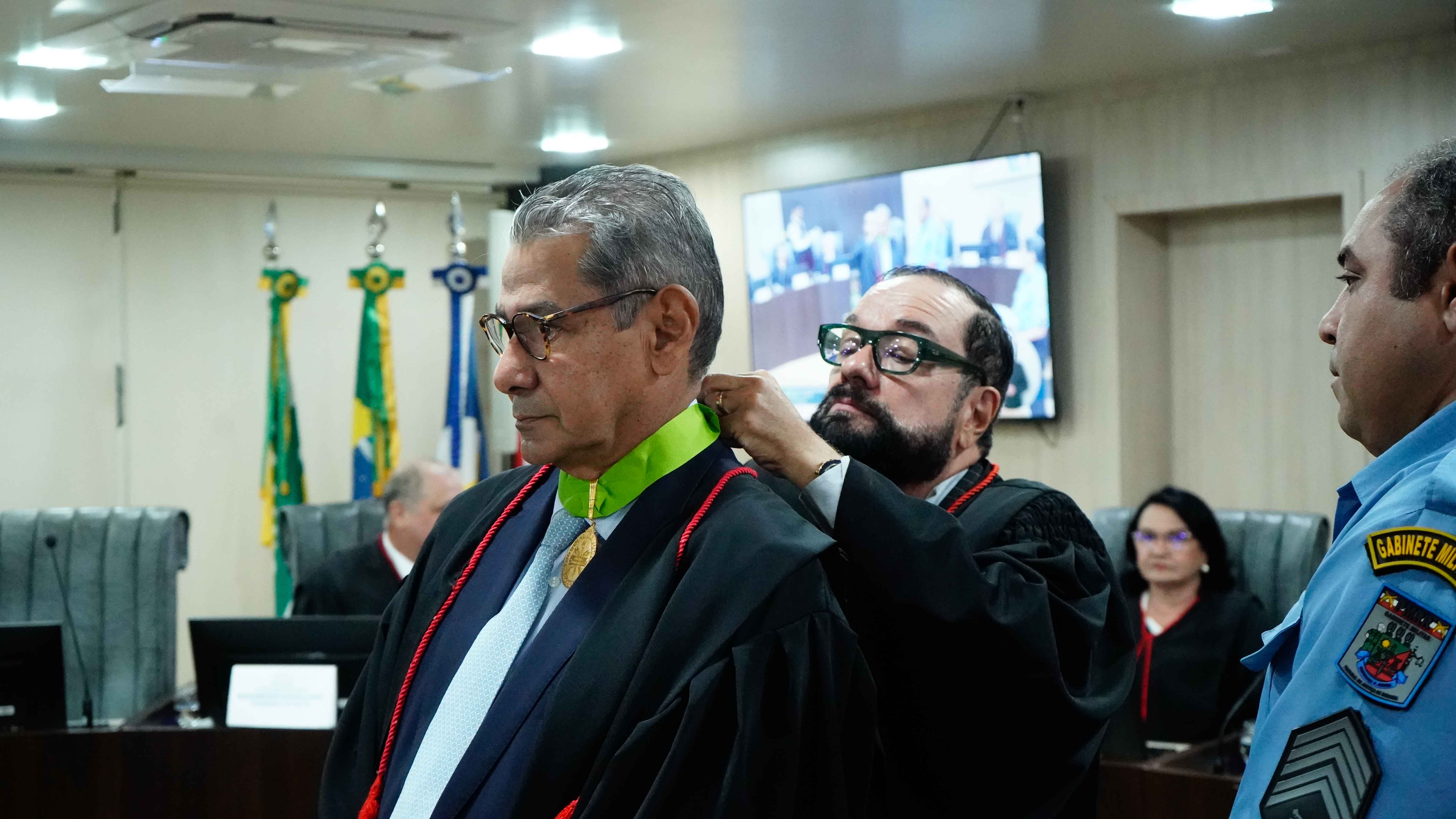 Imagem colorida mostra em cerimônia solene, o presidente do TJRR, Desembargador Leonardo Cupello, ajusta um colar de comenda no Procurador de Justiça Sales Eurico Melgarejo Freitas. Ambos estão de toga em um plenário. Imagem colorida mostra em cerimônia solene, o presidente do TJRR, Desembargador Leonardo Cupello, ajusta um colar de comenda no Procurador de Justiça Sales Eurico Melgarejo Freitas. Ambos estão de toga em um plenário.