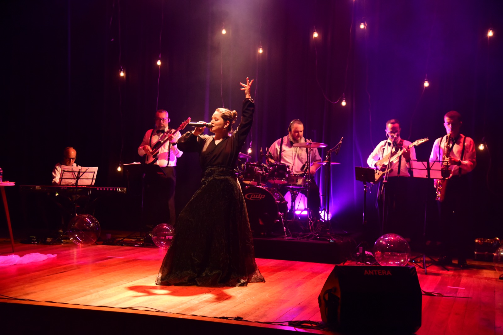 A imagem mostra uma uma cantora em um palco de madeira, sob luzes roxas e amarelas. Ela está ao centro, de vestido preto, cantando ao microfone com o braço direito erguido. Ao fundo, cinco músicos a acompanham tocando bateria, teclado, baixo e guitarras. A imagem mostra uma uma cantora em um palco de madeira, sob luzes roxas e amarelas. Ela está ao centro, de vestido preto, cantando ao microfone com o braço direito erguido. Ao fundo, cinco músicos a acompanham tocando bateria, teclado, baixo e guitarras.