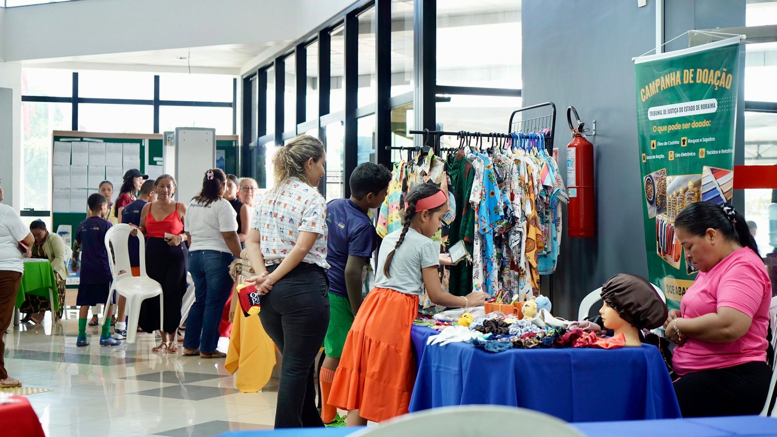 Em um ambiente interno iluminado, várias pessoas circulam entre mesas e estandes organizados em uma feira. À direita, há uma mesa coberta com toalha azul, onde uma mulher de blusa rosa trabalha com tecidos e acessórios expostos. Ao lado, crianças e adultos observam roupas coloridas penduradas em um suporte metálico. Um banner verde com o título “Campanha de Doação” indica que o espaço pertence ao Tribunal de Justiça do Estado de Roraima, informando os tipos de itens que podem ser doados. Ao fundo, mais pessoas interagem e participam da atividade, em um ambiente movimentado e colaborativo.