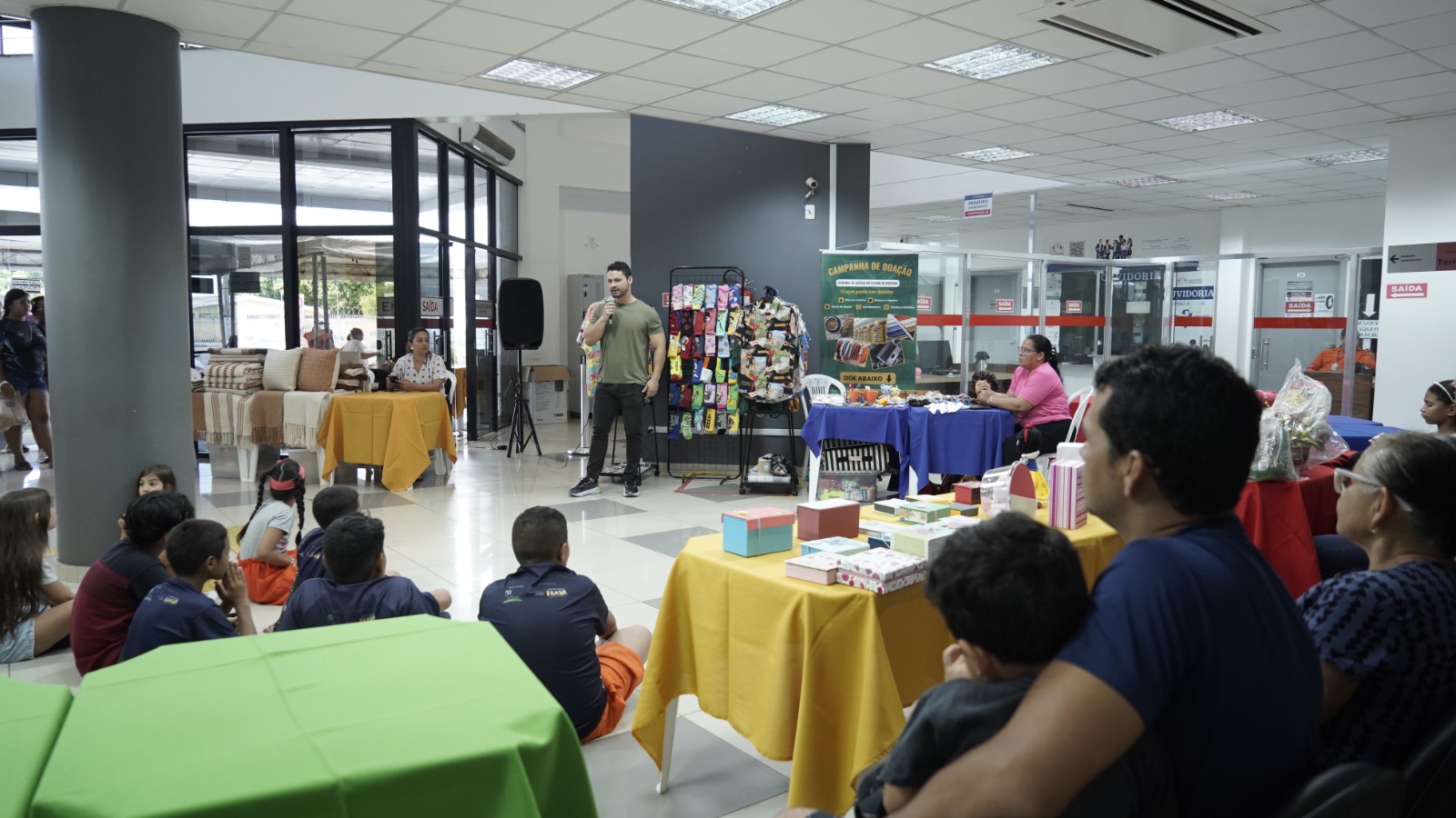 Em um salão amplo e bem iluminado, diversas pessoas participam de uma atividade organizada em formato de feira. No centro da imagem, um homem de camiseta verde fala ao microfone diante de um grupo de crianças sentadas no chão, que o observam atentamente. À direita, há mesas cobertas com toalhas coloridas, onde estão expostos produtos artesanais e caixas decoradas. Atrás do palestrante, vê-se um painel com roupas e um banner verde da “Campanha de Doação” do Tribunal de Justiça do Estado de Roraima. Algumas pessoas estão sentadas ao redor, acompanhando o momento em um ambiente acolhedor e participativo.