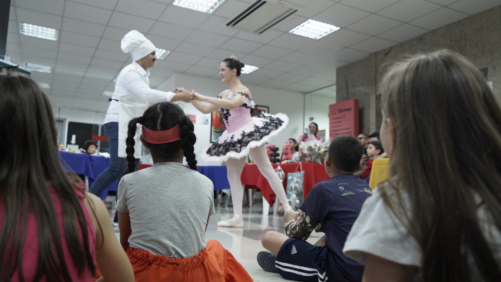 Em um salão iluminado, uma bailarina com figurino rosa e preto dança ao lado de uma pessoa vestida de chef de cozinha, que usa chapéu branco e bigode postiço, formando uma cena divertida. À frente, várias crianças sentadas no chão observam atentamente a apresentação, com expressões de encantamento. Ao fundo, há mesas cobertas com toalhas coloridas e arranjos decorativos, compondo um ambiente alegre e cultural.