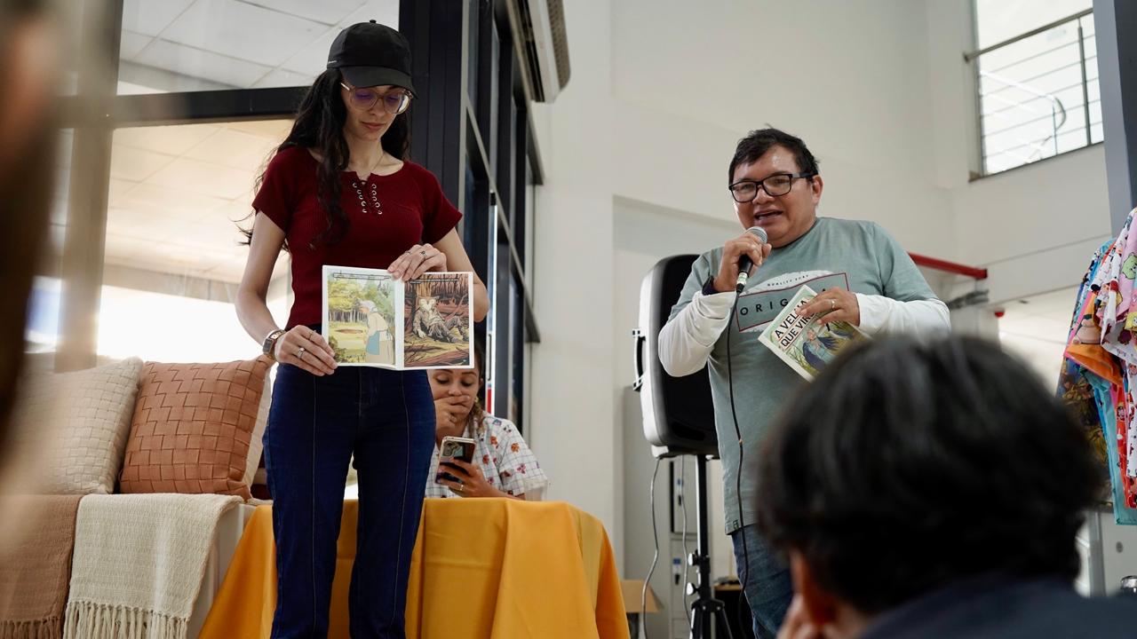 m um ambiente interno, um homem fala ao microfone segurando um livro aberto, enquanto uma jovem ao lado, usando boné preto, óculos e blusa vermelha, exibe as páginas ilustradas da obra para o público. Atrás deles, uma mulher sentada à mesa observa a apresentação e manuseia o celular. O cenário inclui almofadas e mantas expostas sobre uma mesa coberta com toalha amarela, compondo um clima acolhedor e educativo, típico de uma atividade cultural ou de leitura compartilhada.