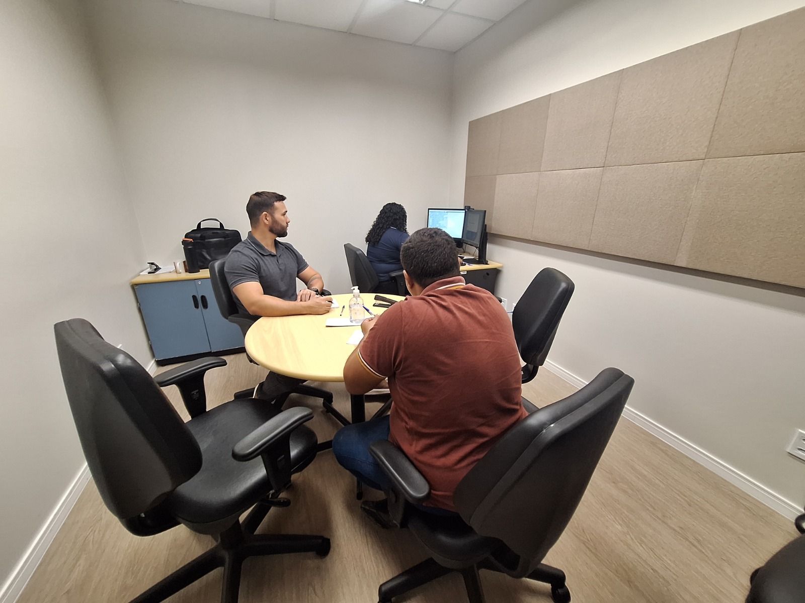 Foto 2: Em uma sala de atendimento, uma mulher de roupa rosa conversa com uma atendente de óculos e casaco azul, que sorri enquanto escreve em um papel. Ao fundo, outra pessoa trabalha em um computador. Sobre a mesa há copos, papéis e um frasco de álcool em gel. O espaço é iluminado e organizado, com cadeiras pretas e paredes claras.