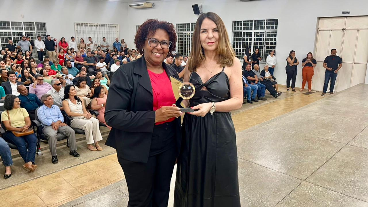 A juíza titular da Comarc Liliane Cardoso ao lado da vereadora Cleudimar Brito em um palco ou frente de sala. A mulher à esquerda, de blazer e óculos, segura um prêmio redondo dourado. Ambas sorriem para a câmera, e ao fundo, um grande público está sentado em cadeiras, assistindo a uma cerimônia ou evento.